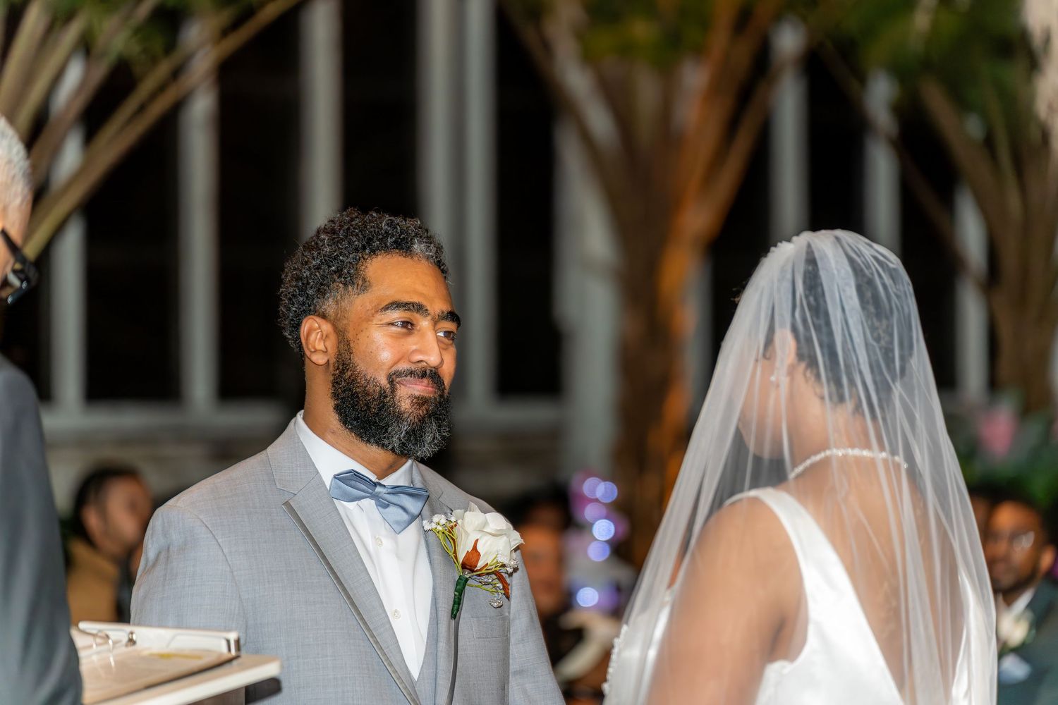 Groom in gray suit with bow tie smiles at bride during outdoor wedding ceremony at night.