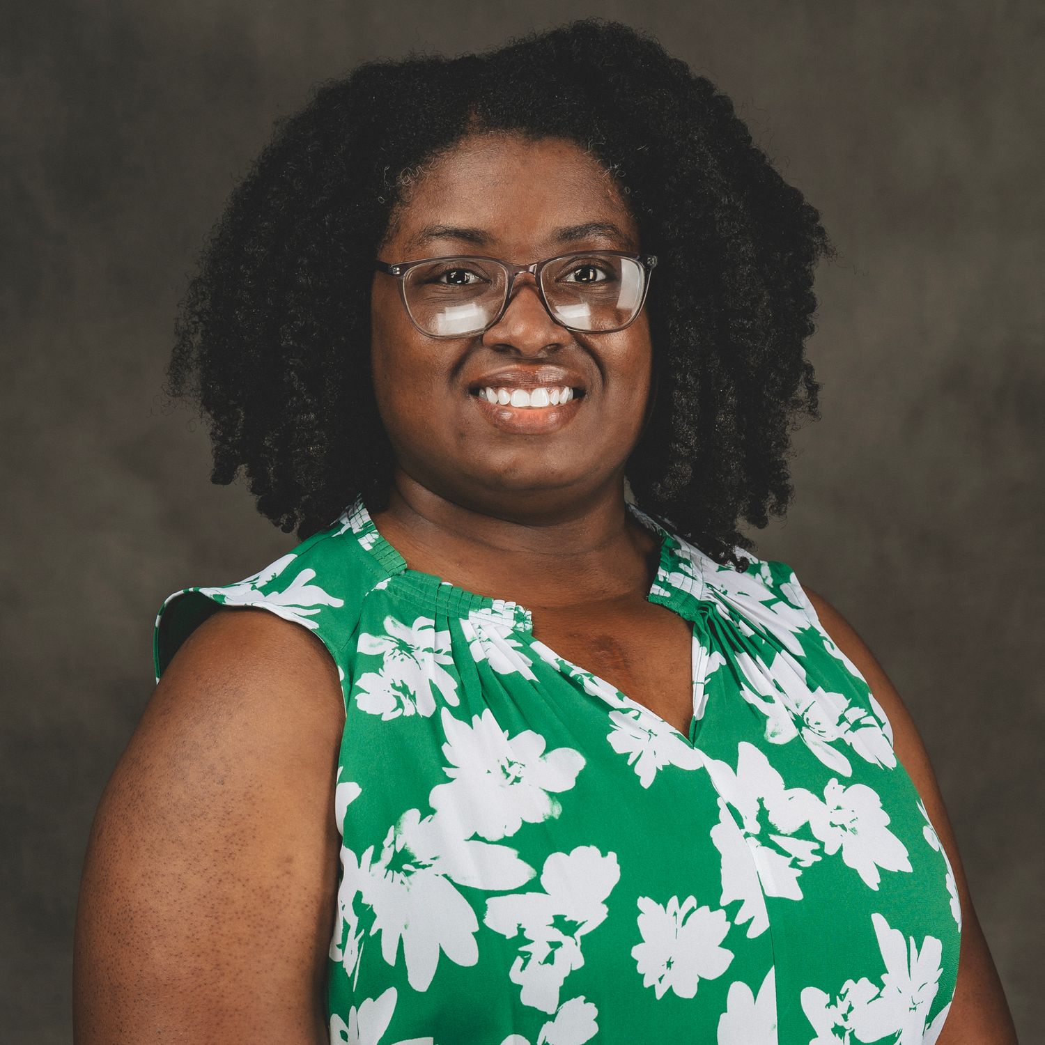 Smiling woman with natural hair and glasses wearing a green floral sleeveless top in a professional headshot.