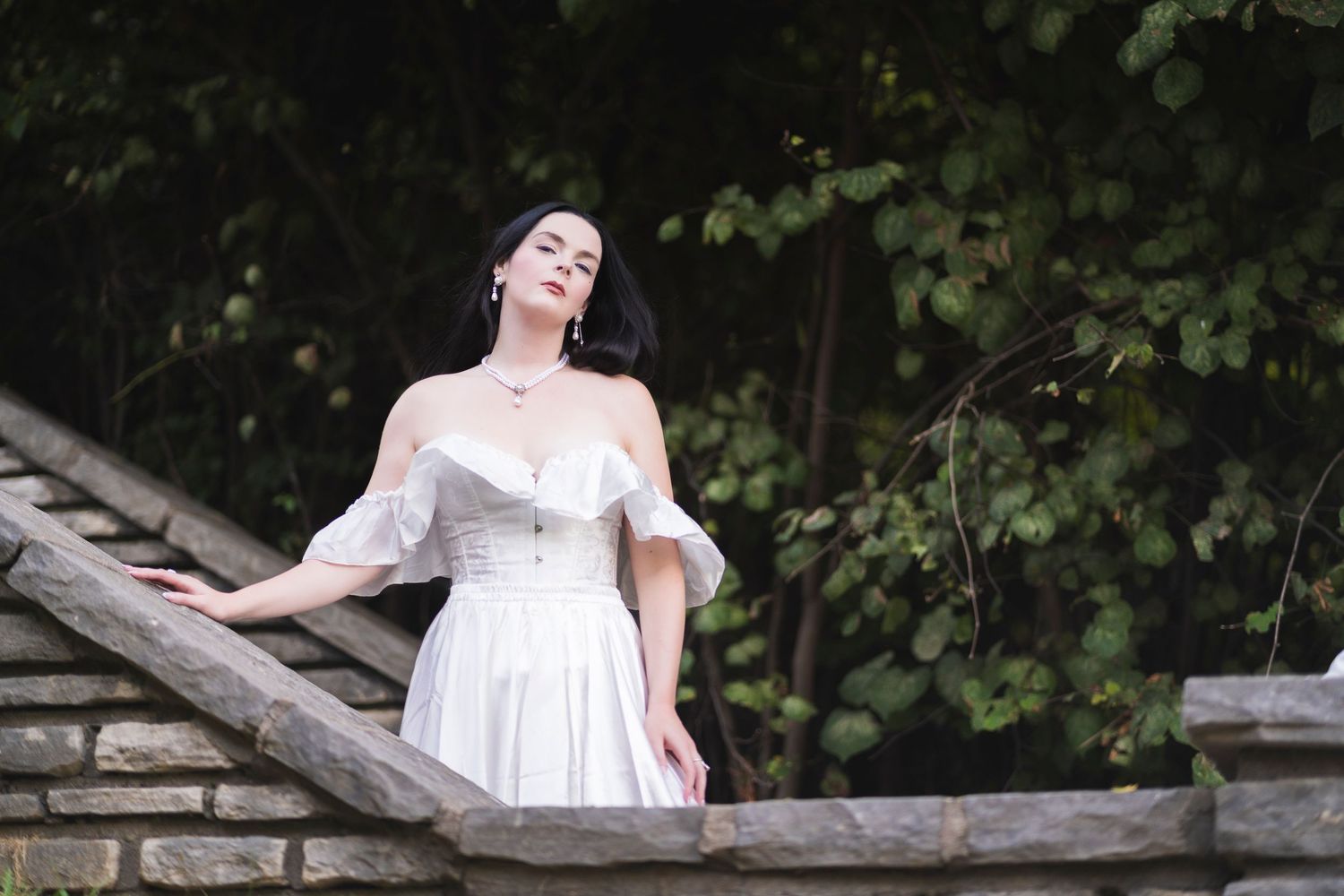 Dark-haired woman in off-shoulder white corset dress on stone steps surrounded by lush greenery.