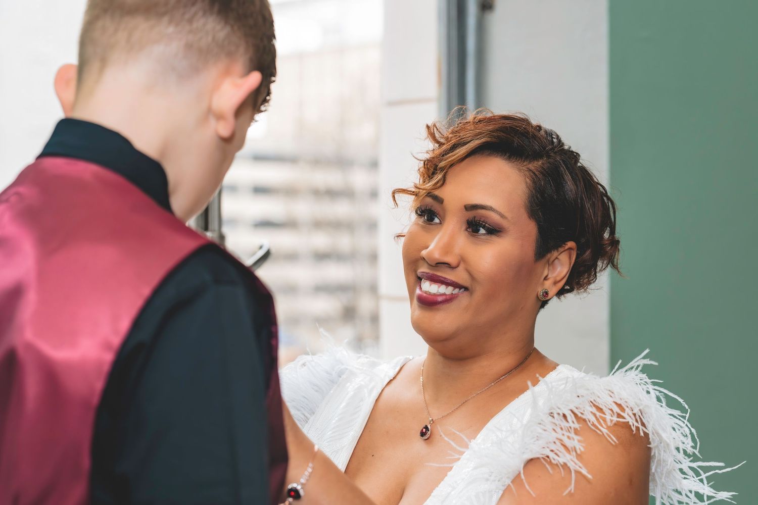 A bride in a white feathered dress smiles warmly at a young man in a burgundy vest.