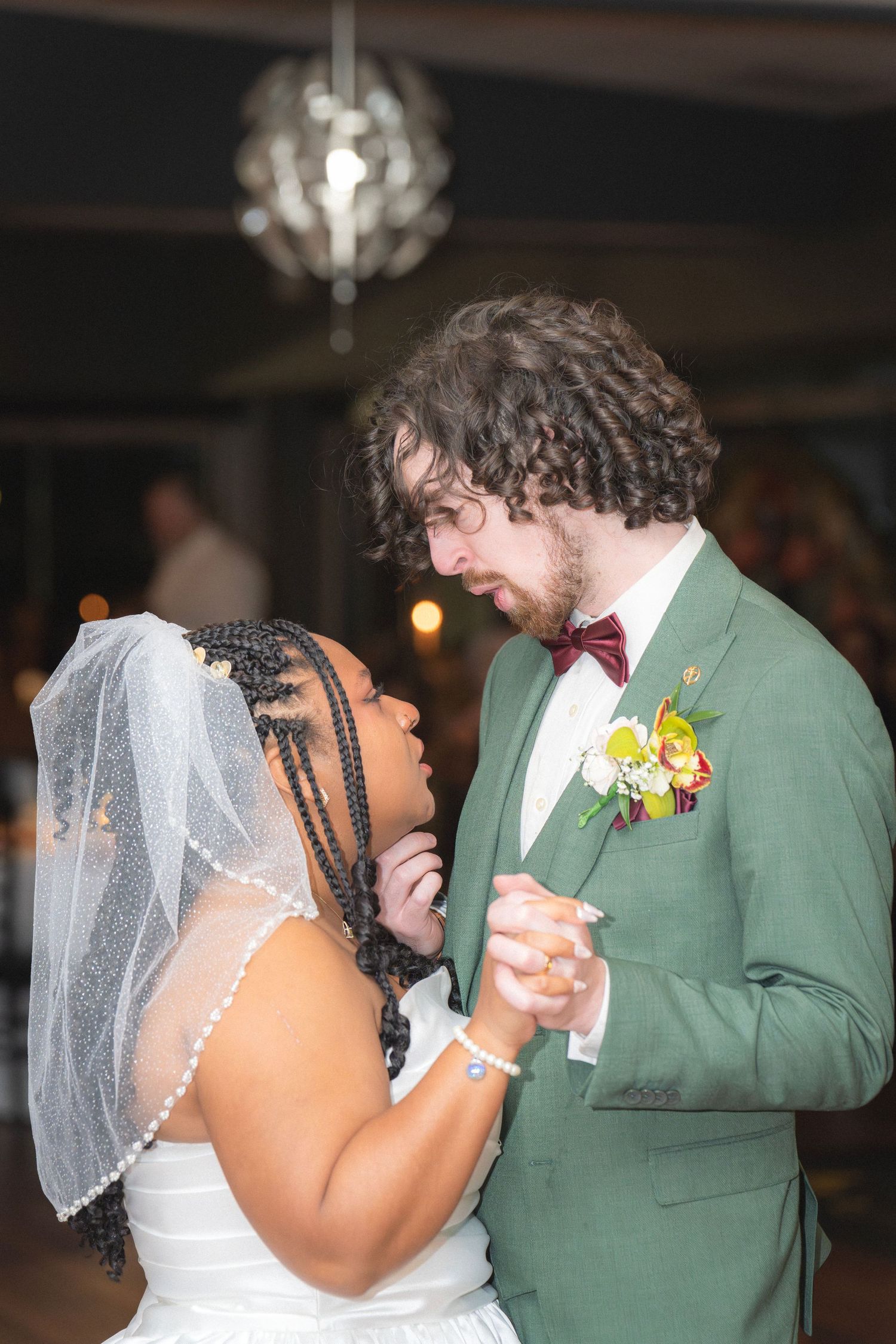 Interracial couple shares first dance, bride in white gown with veil, groom in green suit with boutonniere.