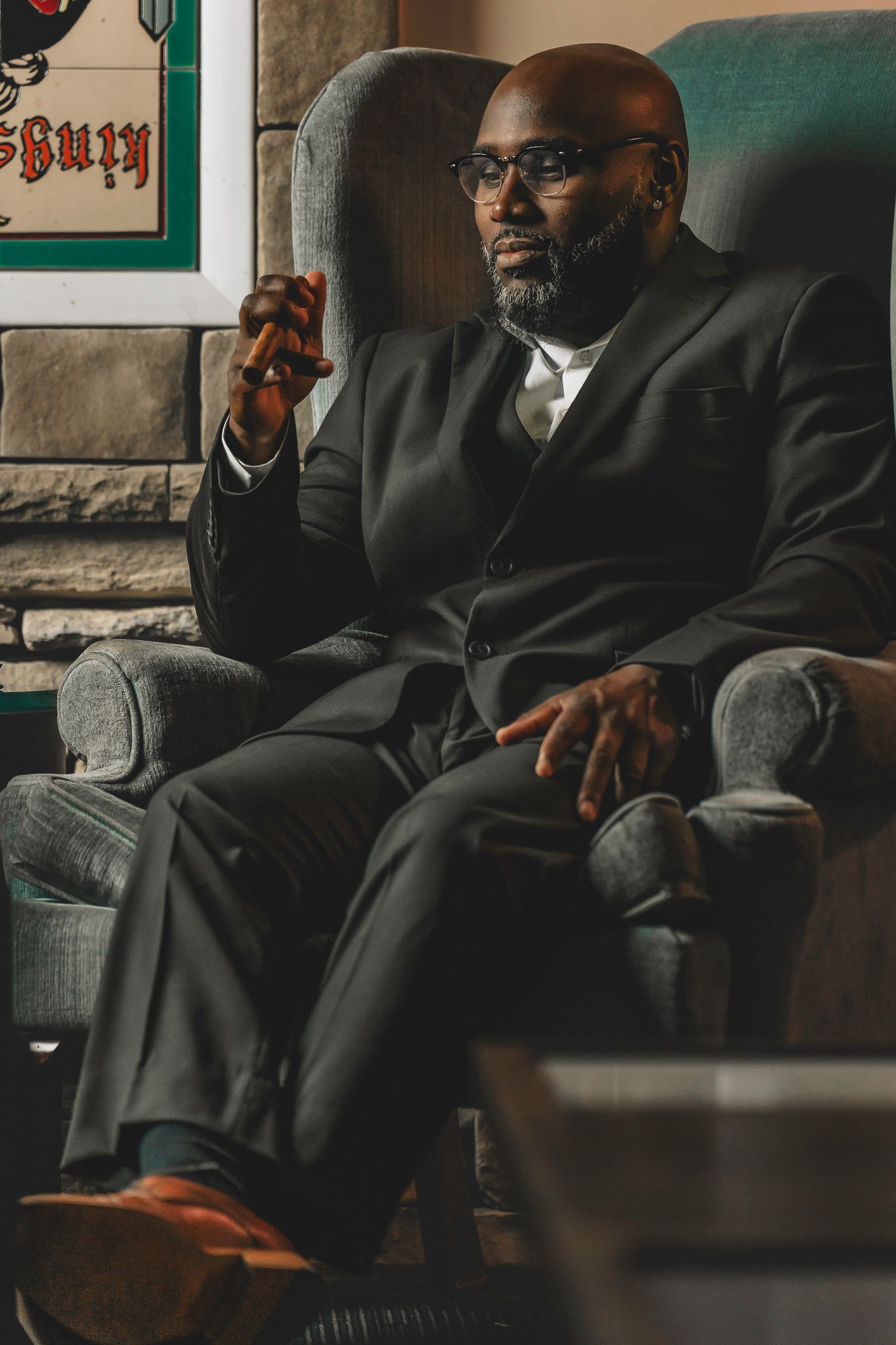 Distinguished man in black suit sitting in armchair holding cigar with confident pose.