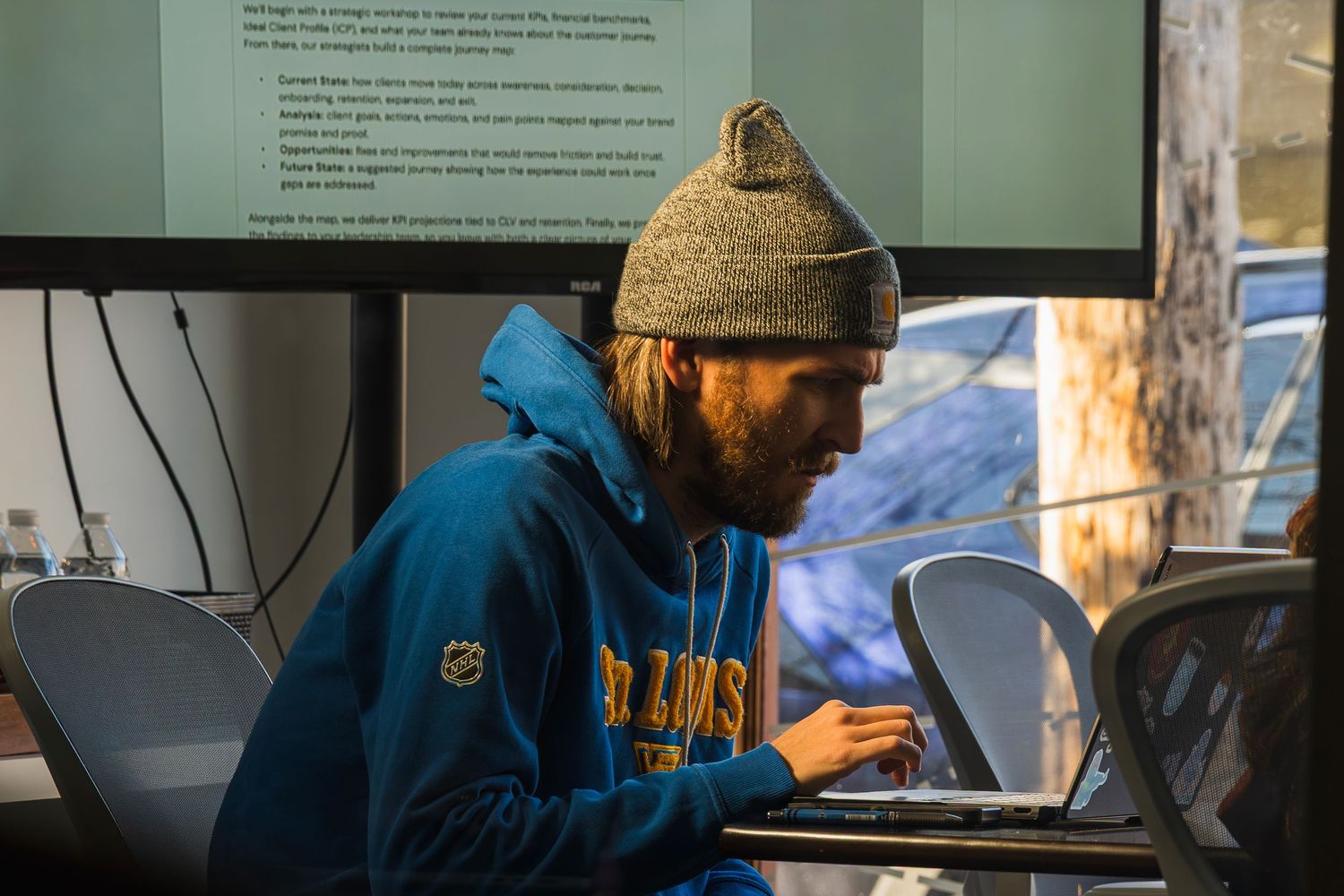 Bearded man in blue hoodie and gray beanie working on laptop in dimly lit workspace.