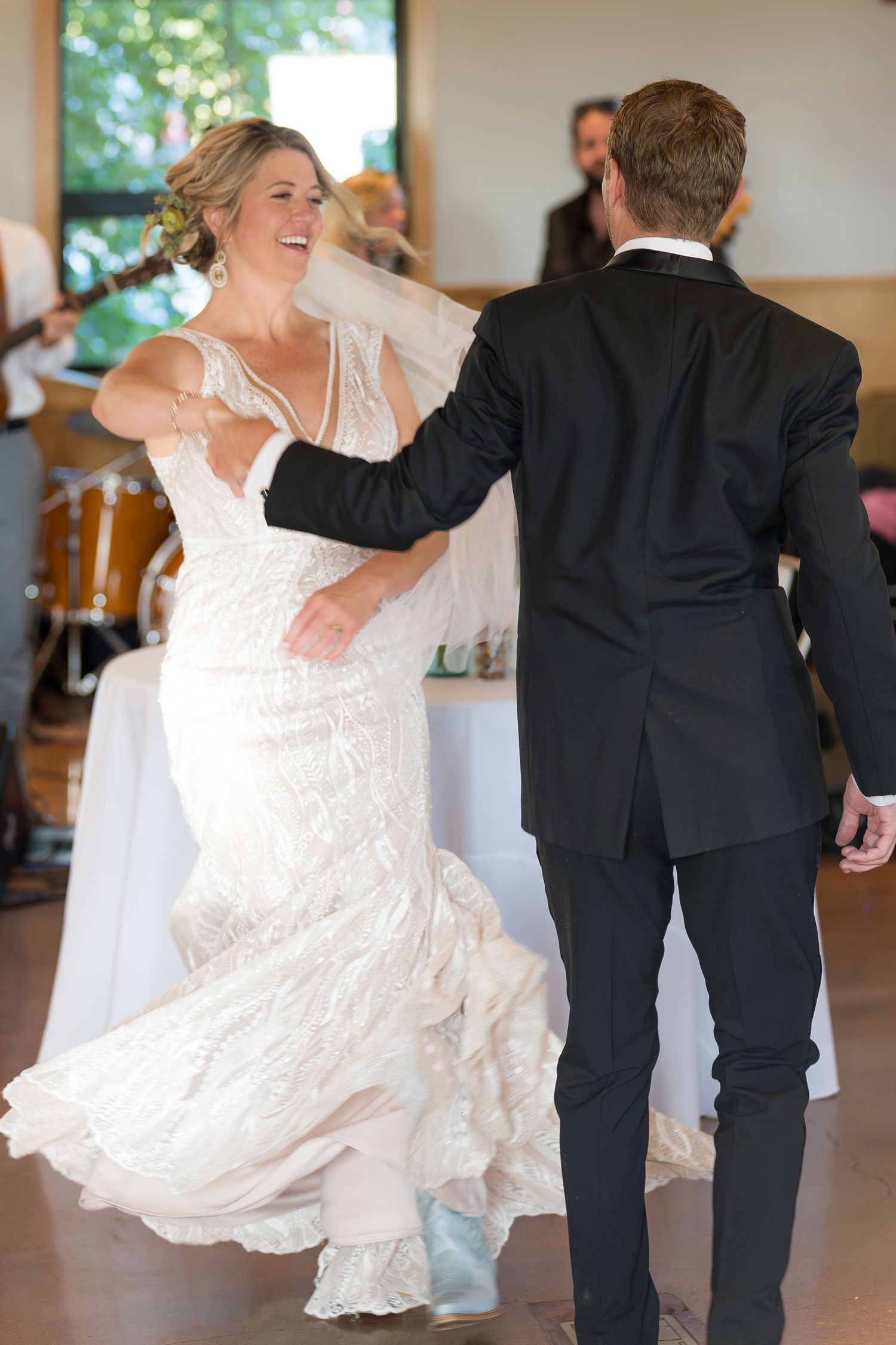 Bride in lace gown twirling with groom in black suit during first dance at wedding reception.