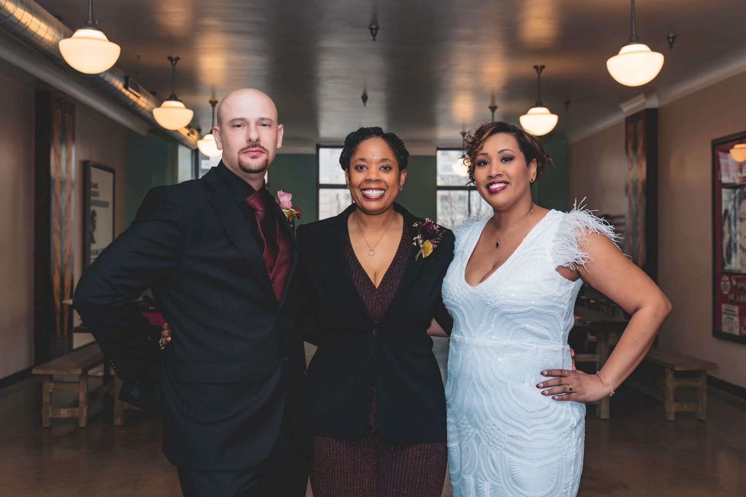 Bride in white feathered gown poses with two guests in a warmly lit wedding venue hallway.