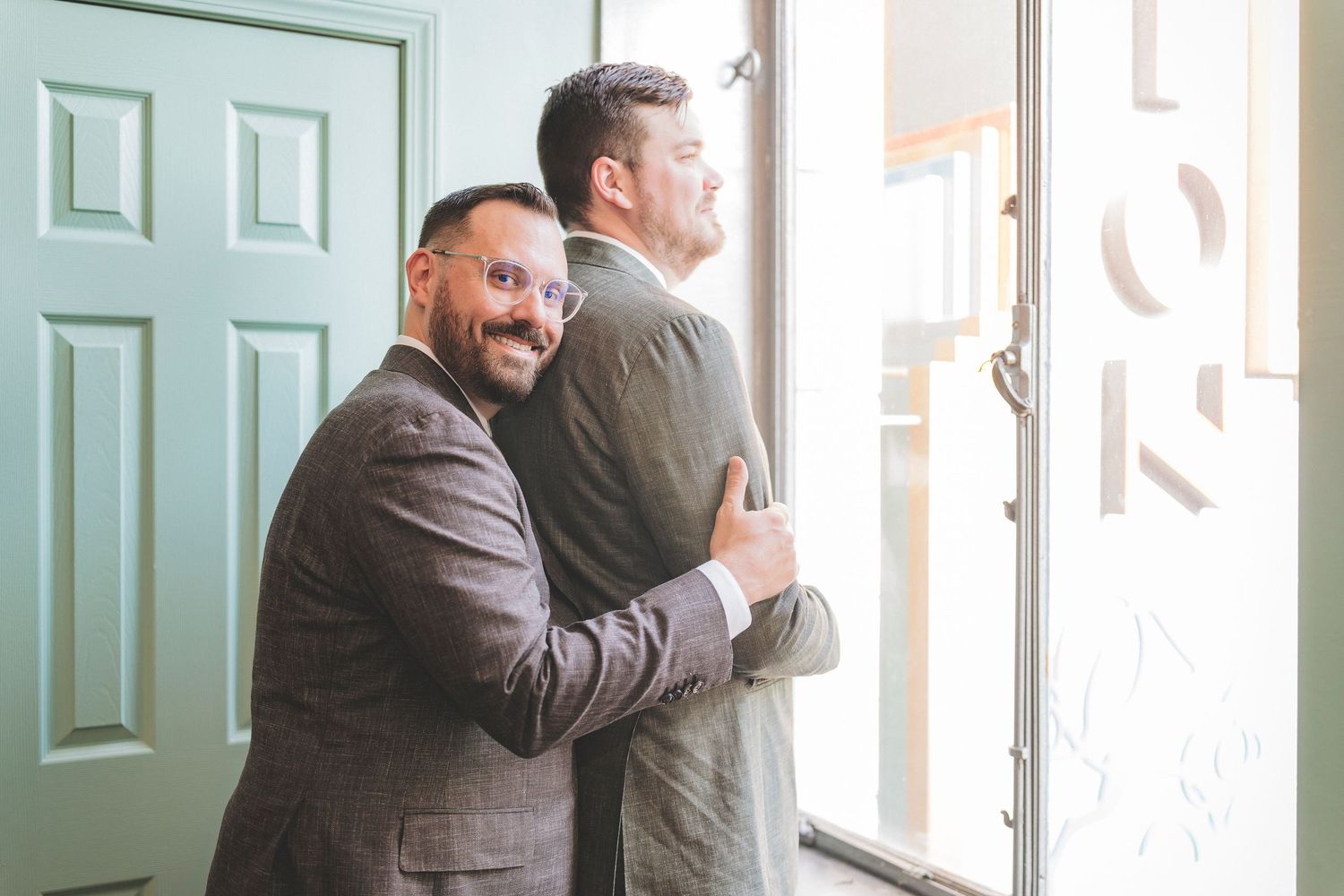 Two grooms in matching gray suits share a warm embrace near a mint green door on their wedding day.