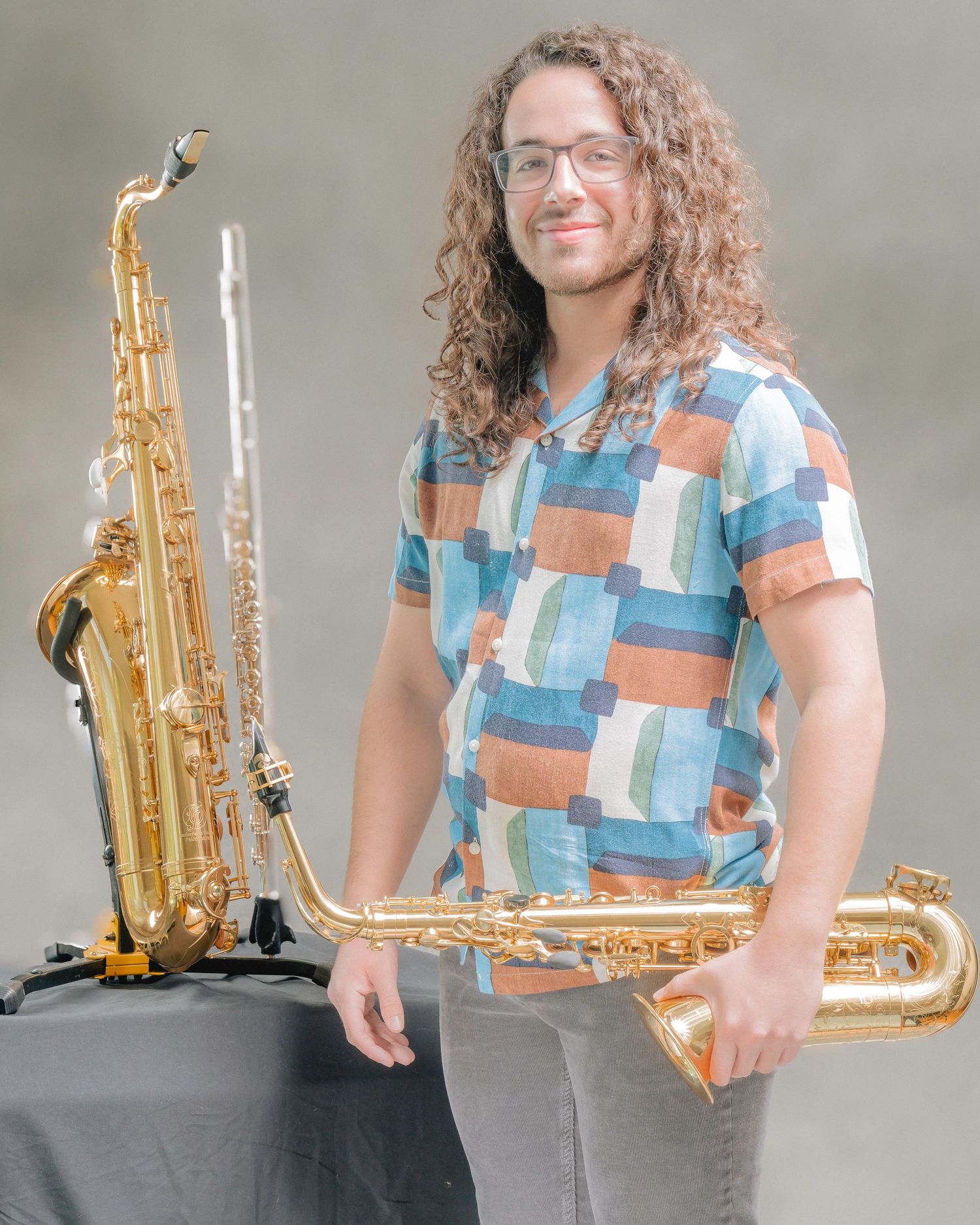 Young man with curly hair holding saxophones outdoors in a colorful patchwork shirt.