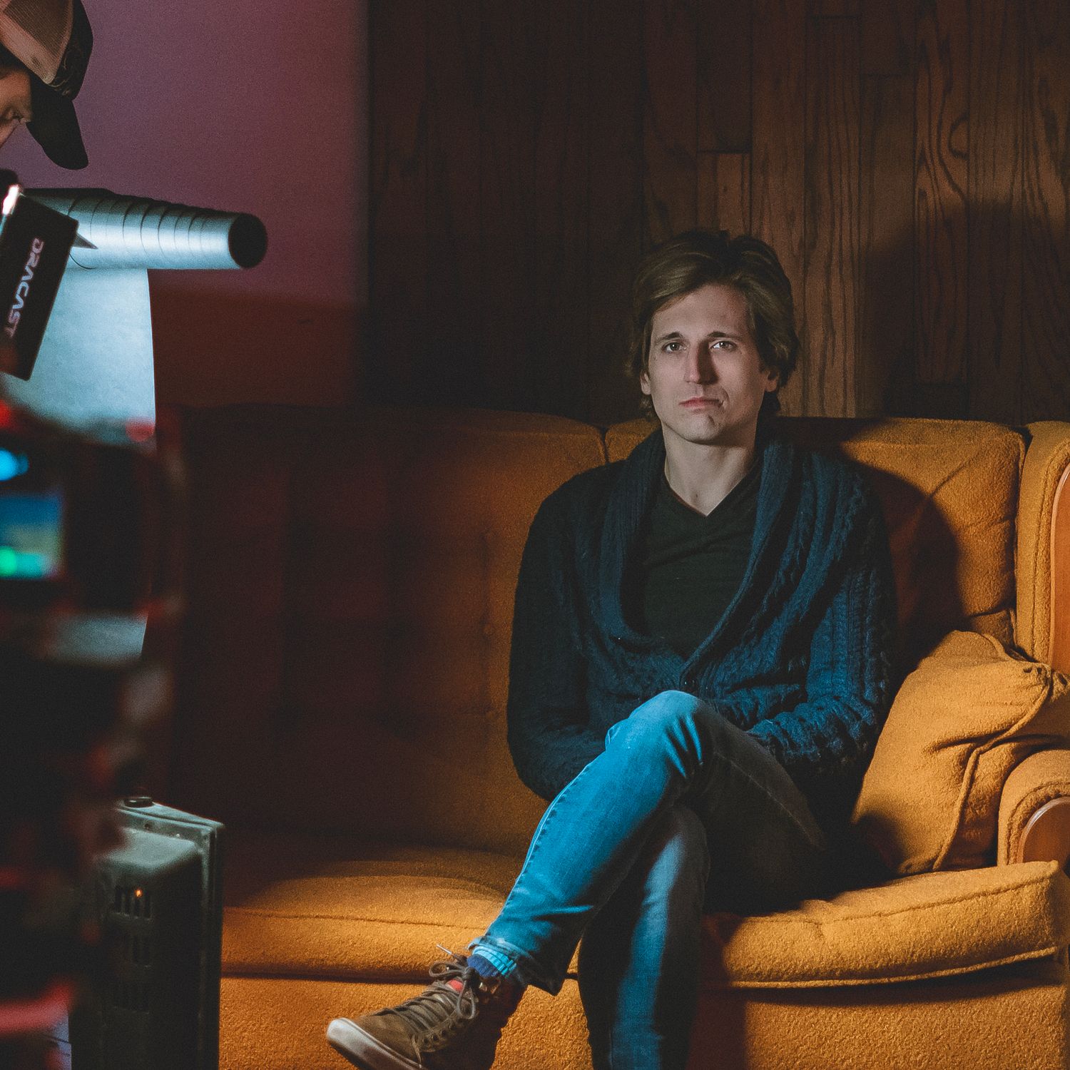Young man in casual clothing sits on a vintage orange sofa being filmed by a professional video camera in dim lighting.
