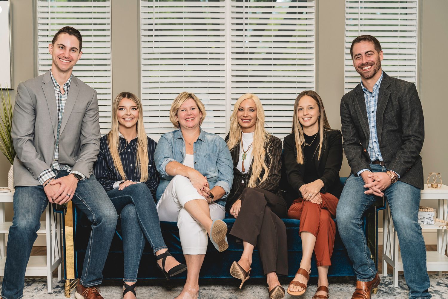 Six smiling professionals pose together on a couch in a bright modern office setting for a team photo.