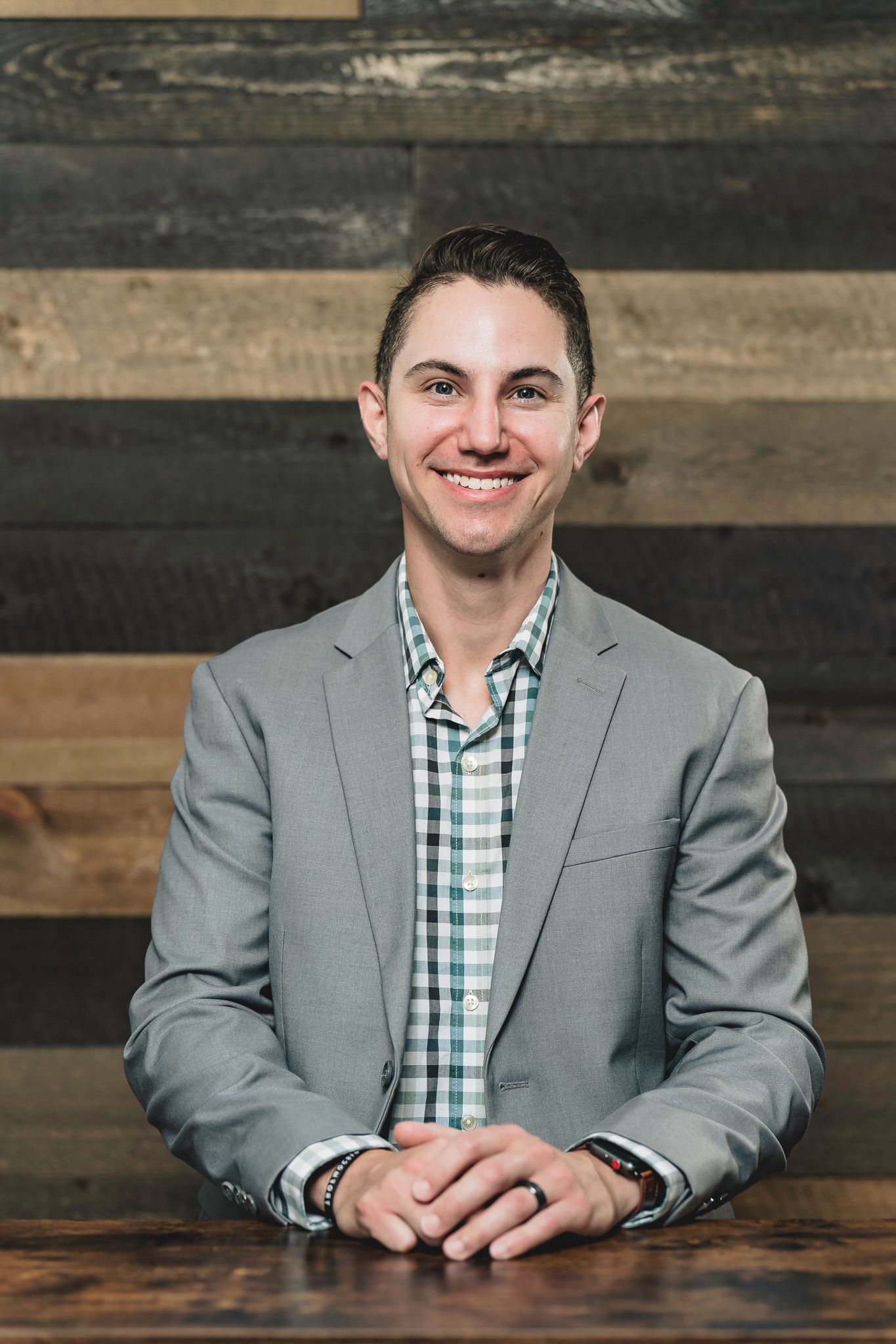 Smiling young professional man in gray blazer and plaid shirt seated at wooden table with rustic wood wall background.