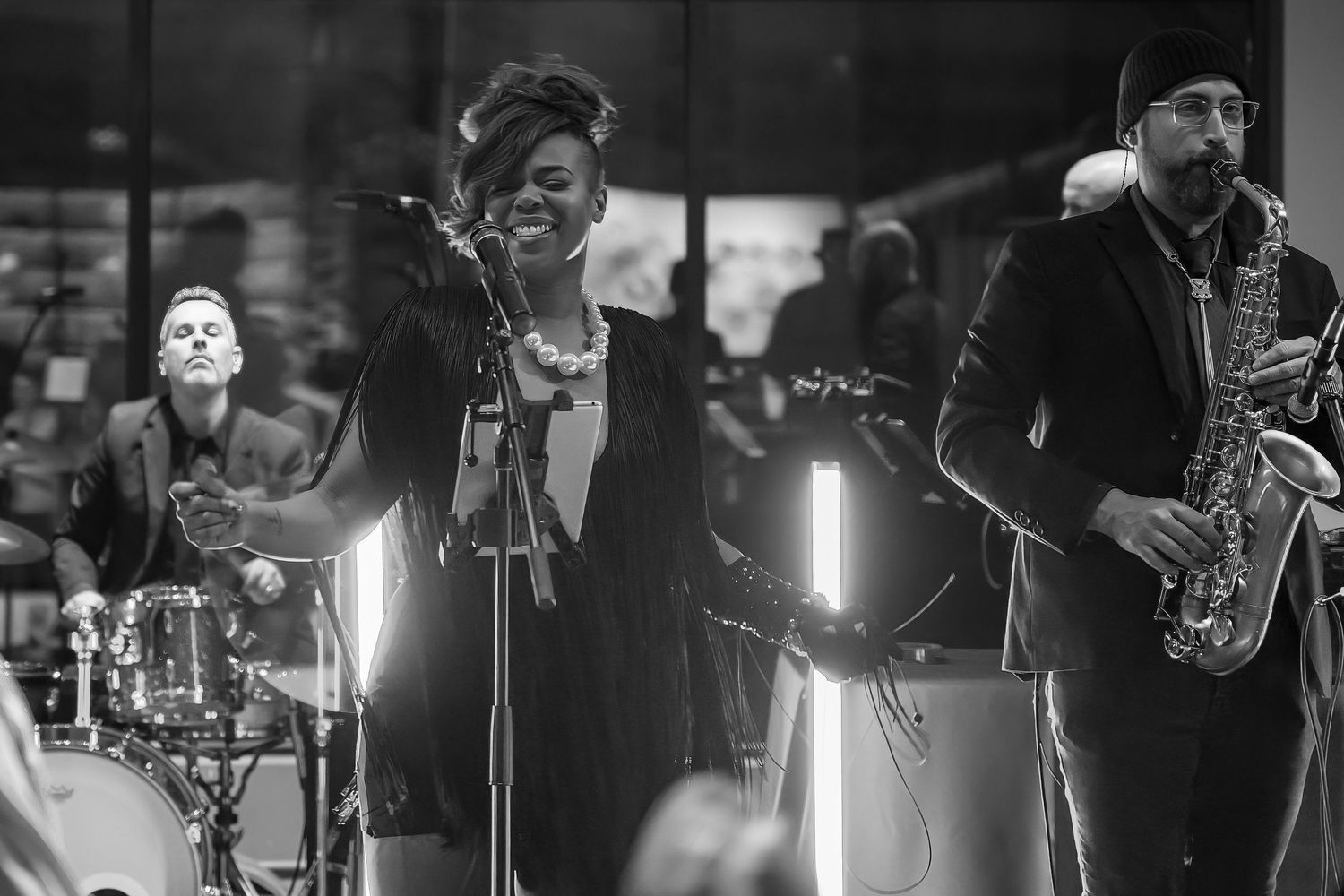 Black and white photo of a female vocalist performing on stage with saxophonist and drummer.