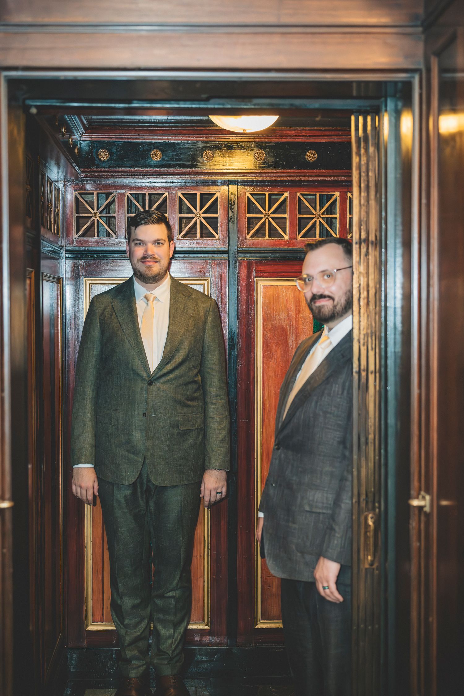 Two grooms in elegant suits pose together inside a vintage wooden elevator for wedding photos.