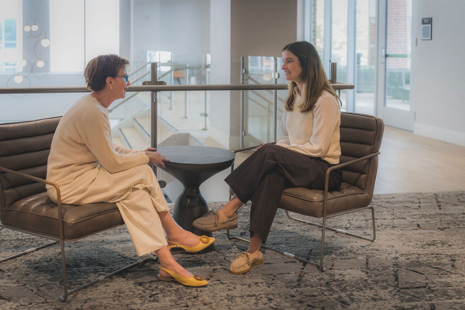 Two professional women having conversation seated in modern office lounge area.