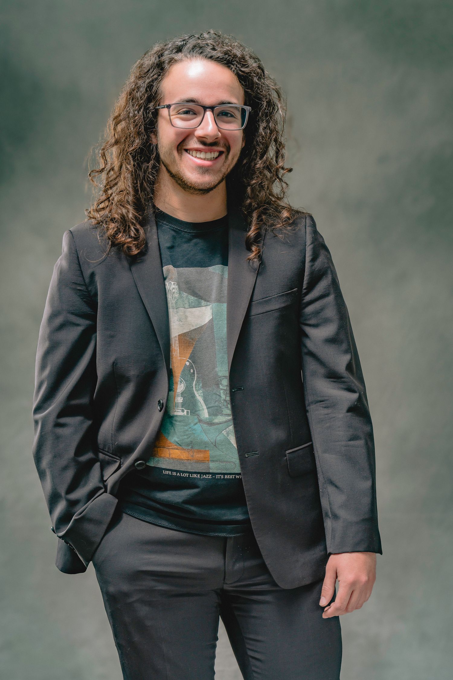 Young man with long curly hair and glasses smiling in a dark blazer over graphic tee.