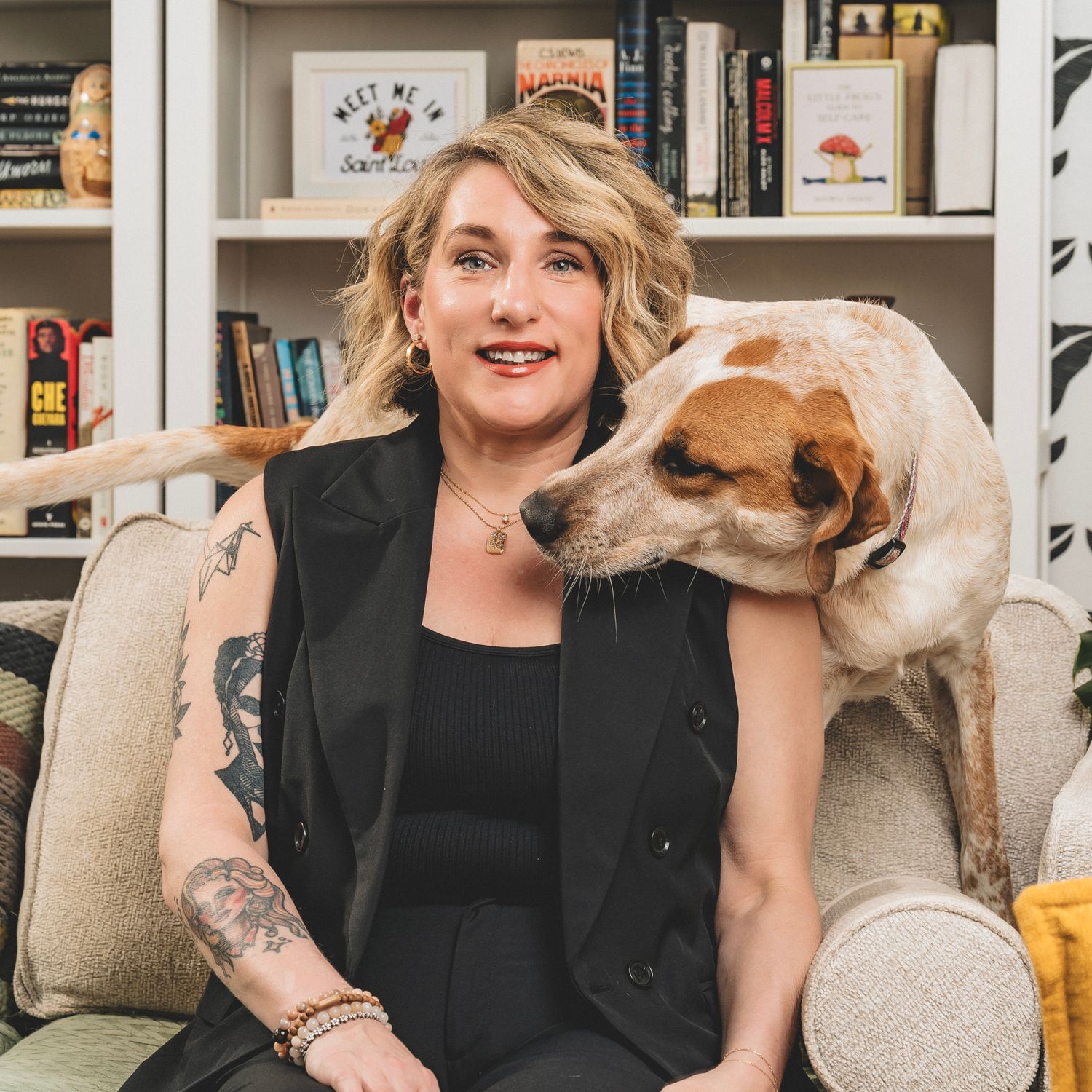 Smiling woman with tattoos sits on a couch with a friendly dog resting its head on her shoulder near a bookshelf.