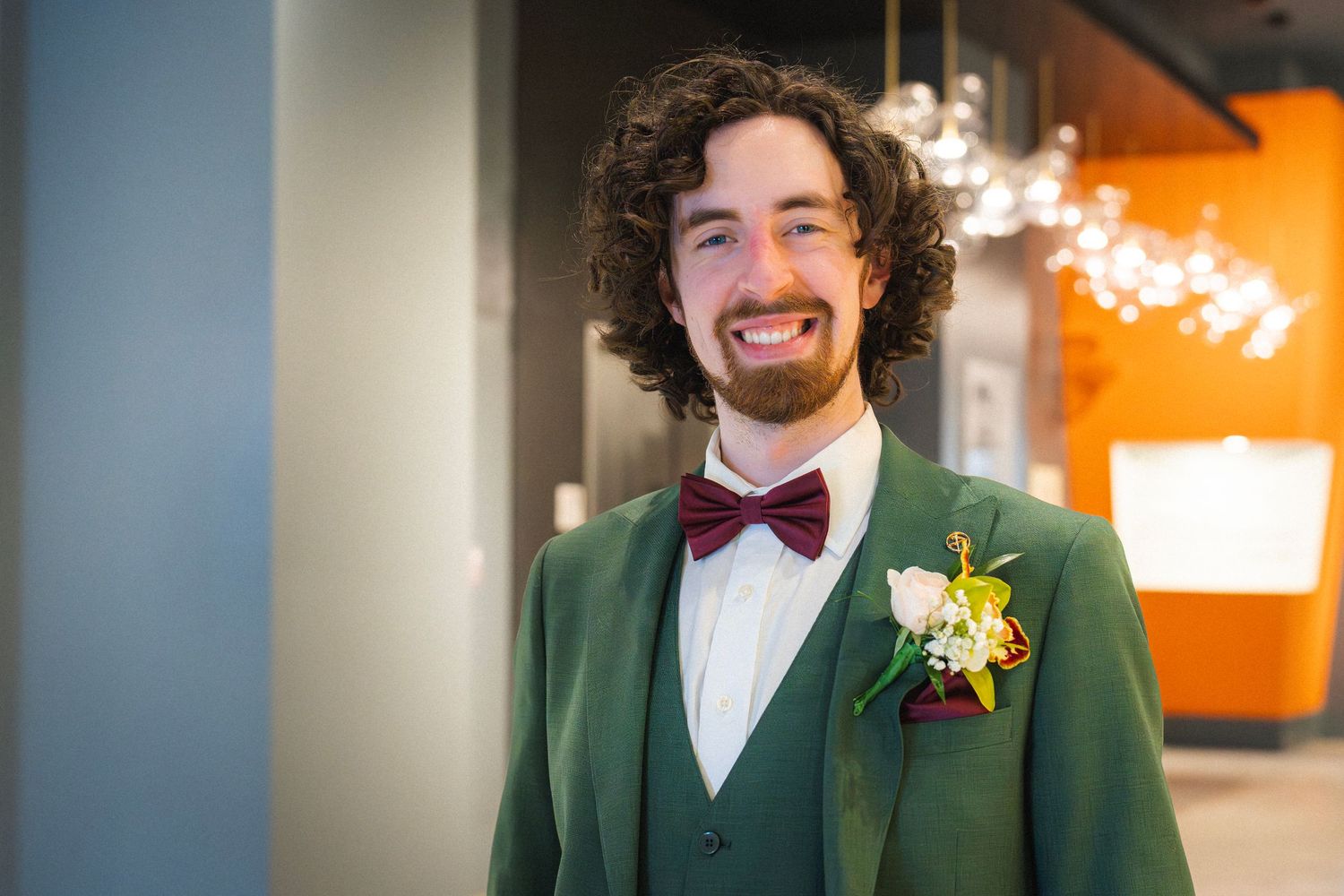 Smiling groom in green suit with burgundy bow tie and floral boutonniere at wedding venue.