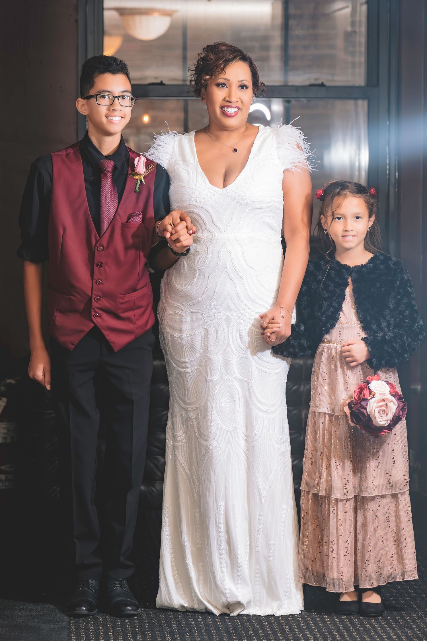 Smiling bride in white feathered gown poses with young boy in burgundy vest and little girl in pink dress.