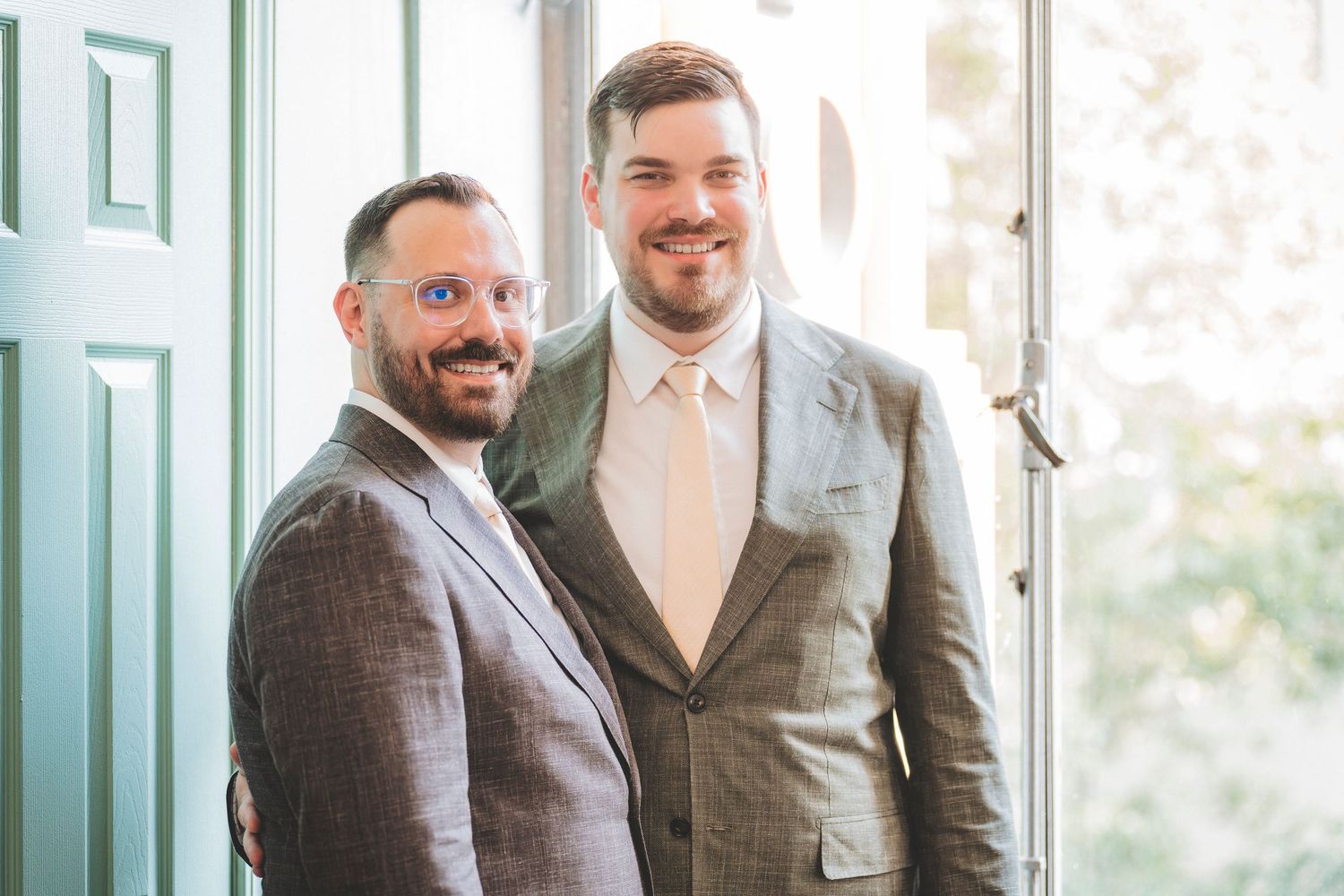 Two grooms in light gray suits pose together smiling near a window at their wedding venue.