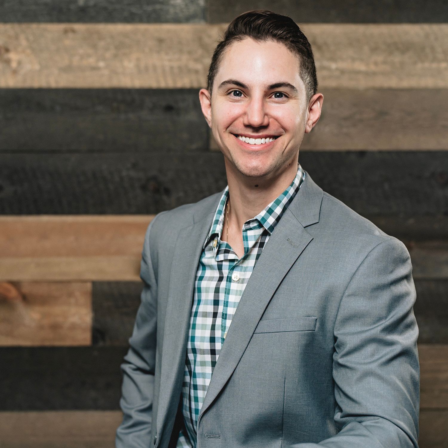 Young smiling man in a gray blazer and plaid shirt posing for a professional headshot.