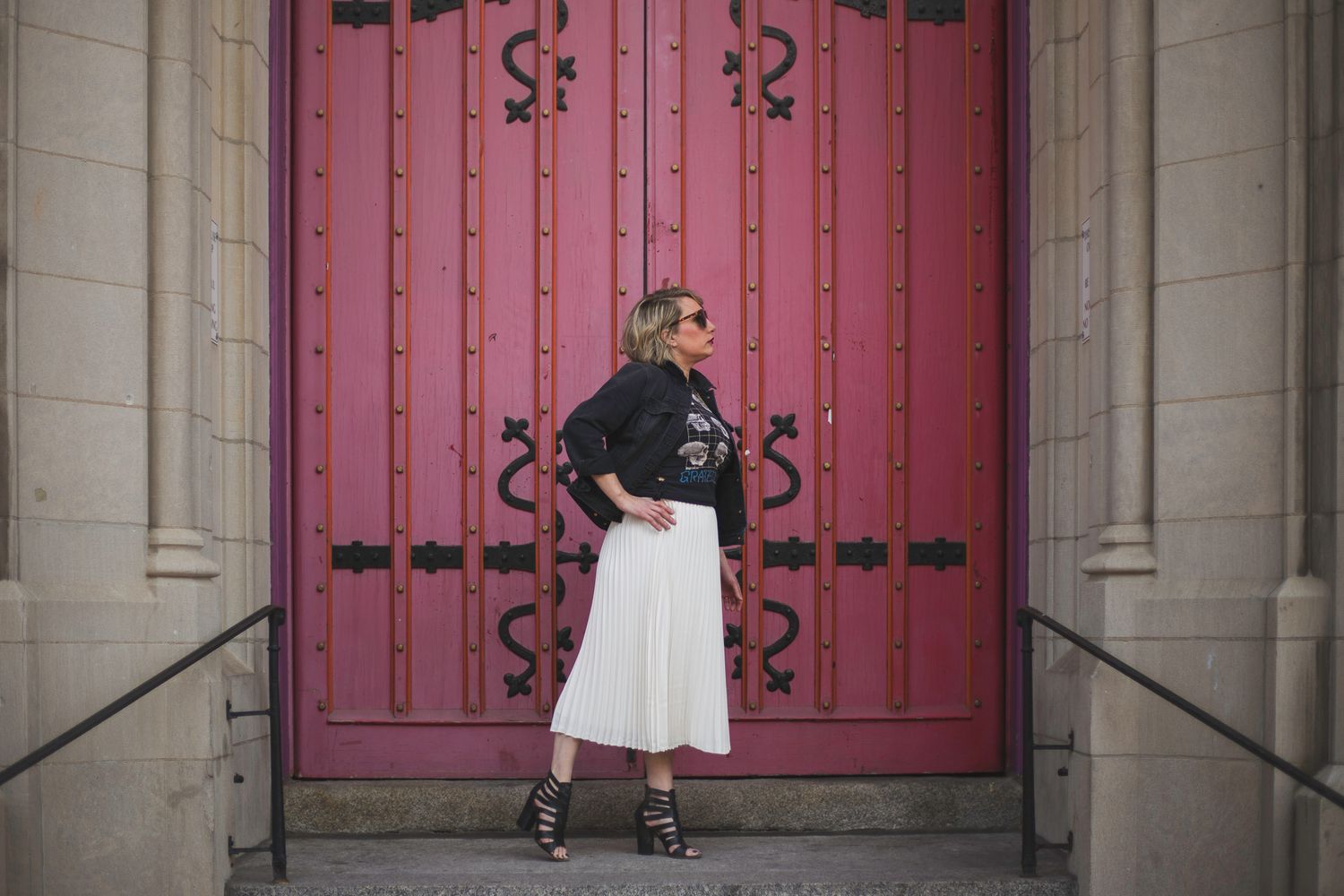 Stylish woman in a leather jacket and white skirt stands before large ornate pink double doors.