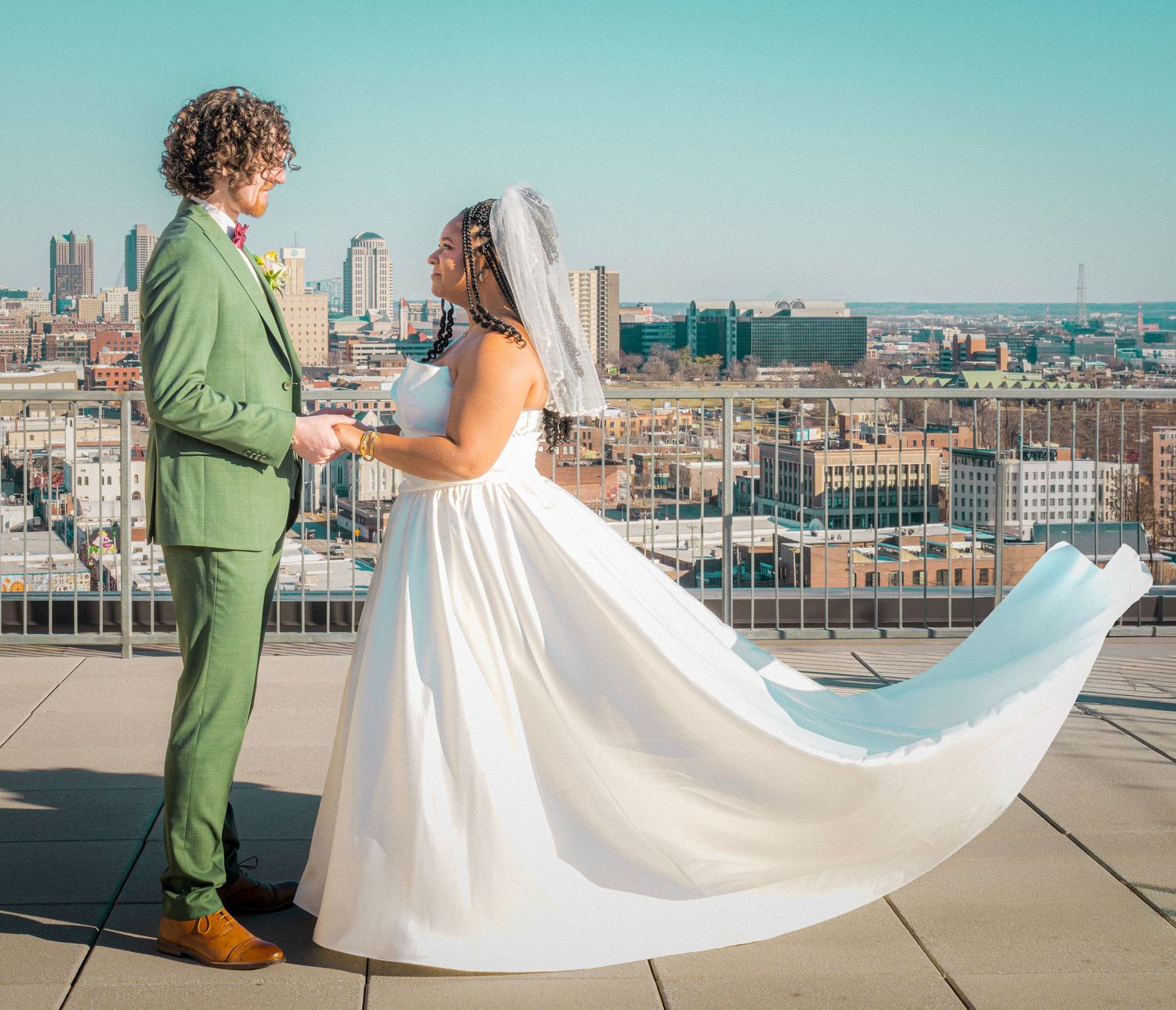 Bride and groom holding hands on rooftop with city skyline backdrop, groom in green suit, bride's dress flowing in wind.