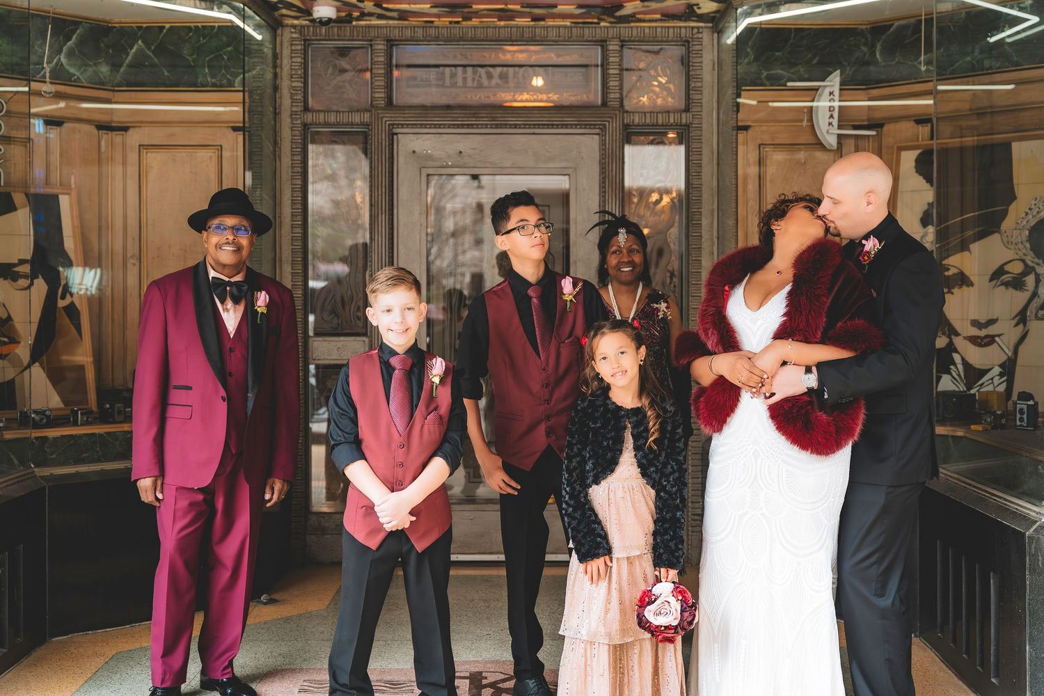Wedding party poses together inside venue with bride in white gown and burgundy fur wrap.