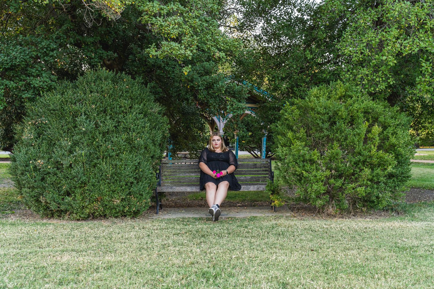 Woman sits alone on a wooden bench surrounded by lush green trees and shrubs in a park setting.