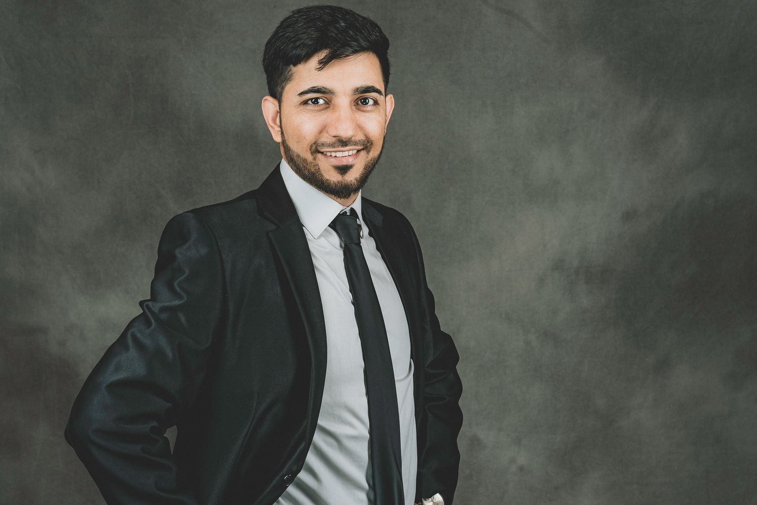 Professional man in a dark suit and tie poses confidently against a gray studio background.