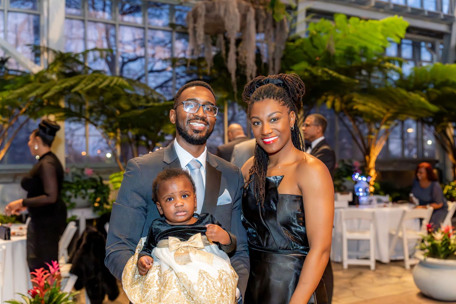 Couple in formal attire holding baby at elegant indoor garden event venue.