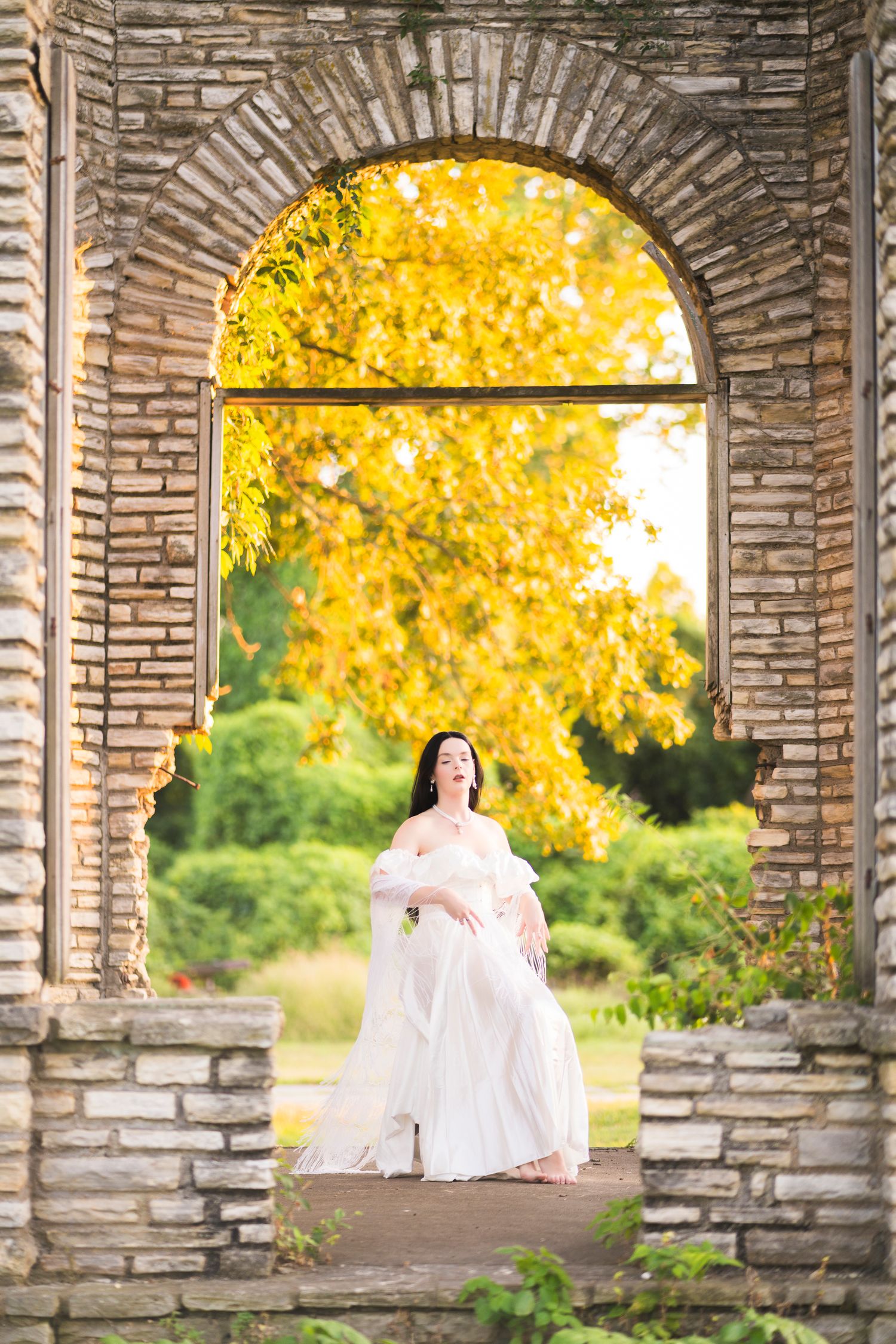 Woman in flowing white dress seated in brick archway with vibrant yellow autumn foliage backdrop.
