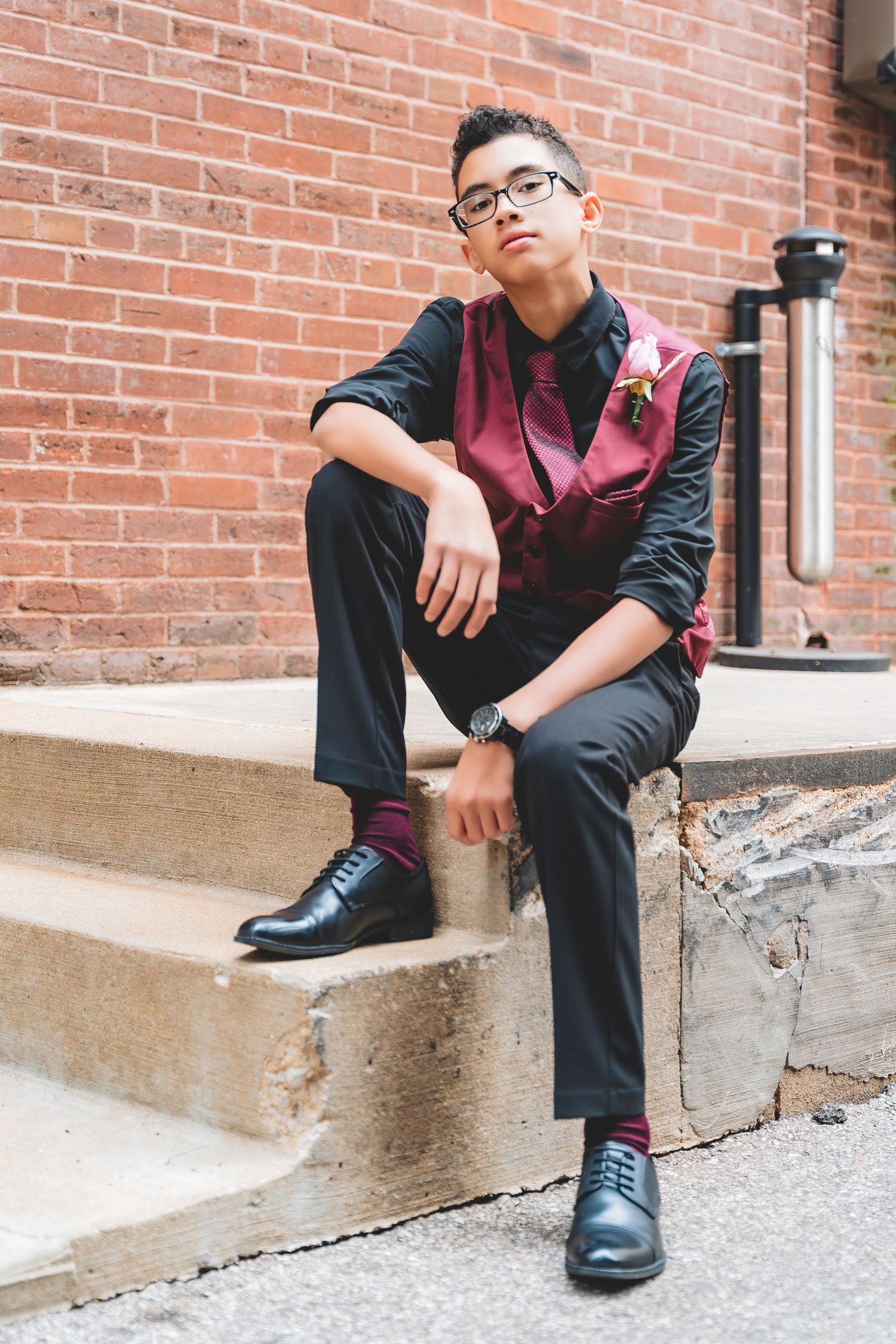 Young teen groomsman in burgundy vest and glasses sits on outdoor brick steps.