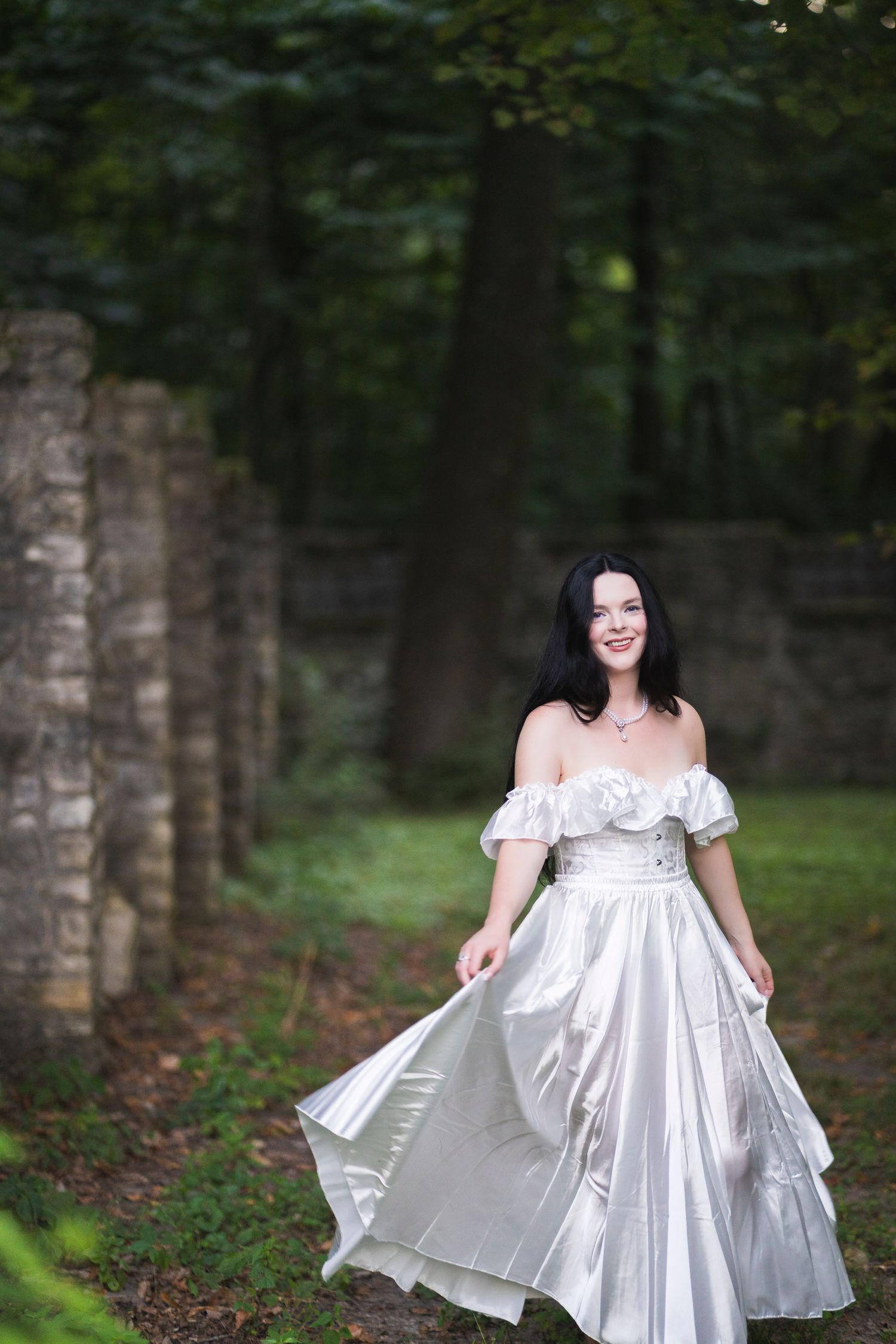 A woman in an elegant white off-shoulder satin gown walks through a lush green forest pathway near stone pillars.
