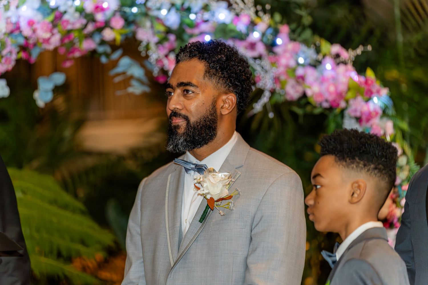 Handsome groom in gray suit with boutonniere standing at wedding altar with young boy.