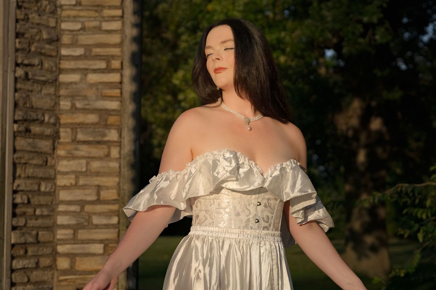 Woman in off-shoulder white satin gown poses outdoors in golden light.
