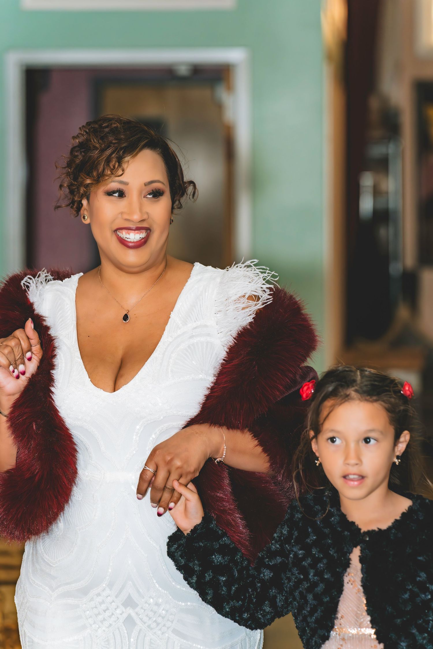 Bride in white gown with burgundy fur shawl holds hands with young flower girl.