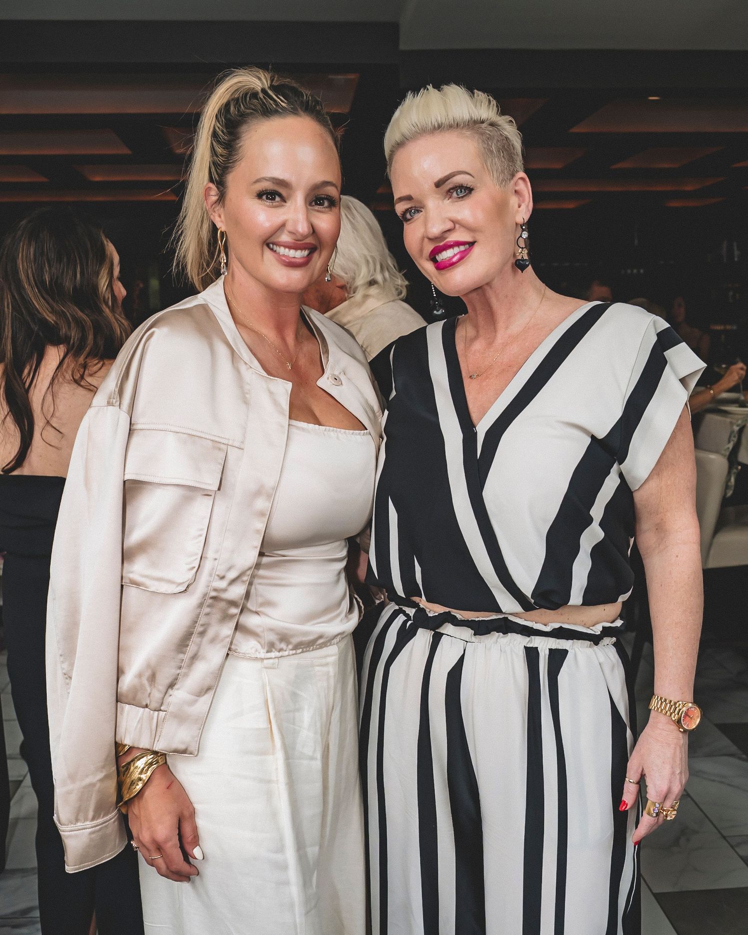 Two stylish women smile together at an indoor social event.