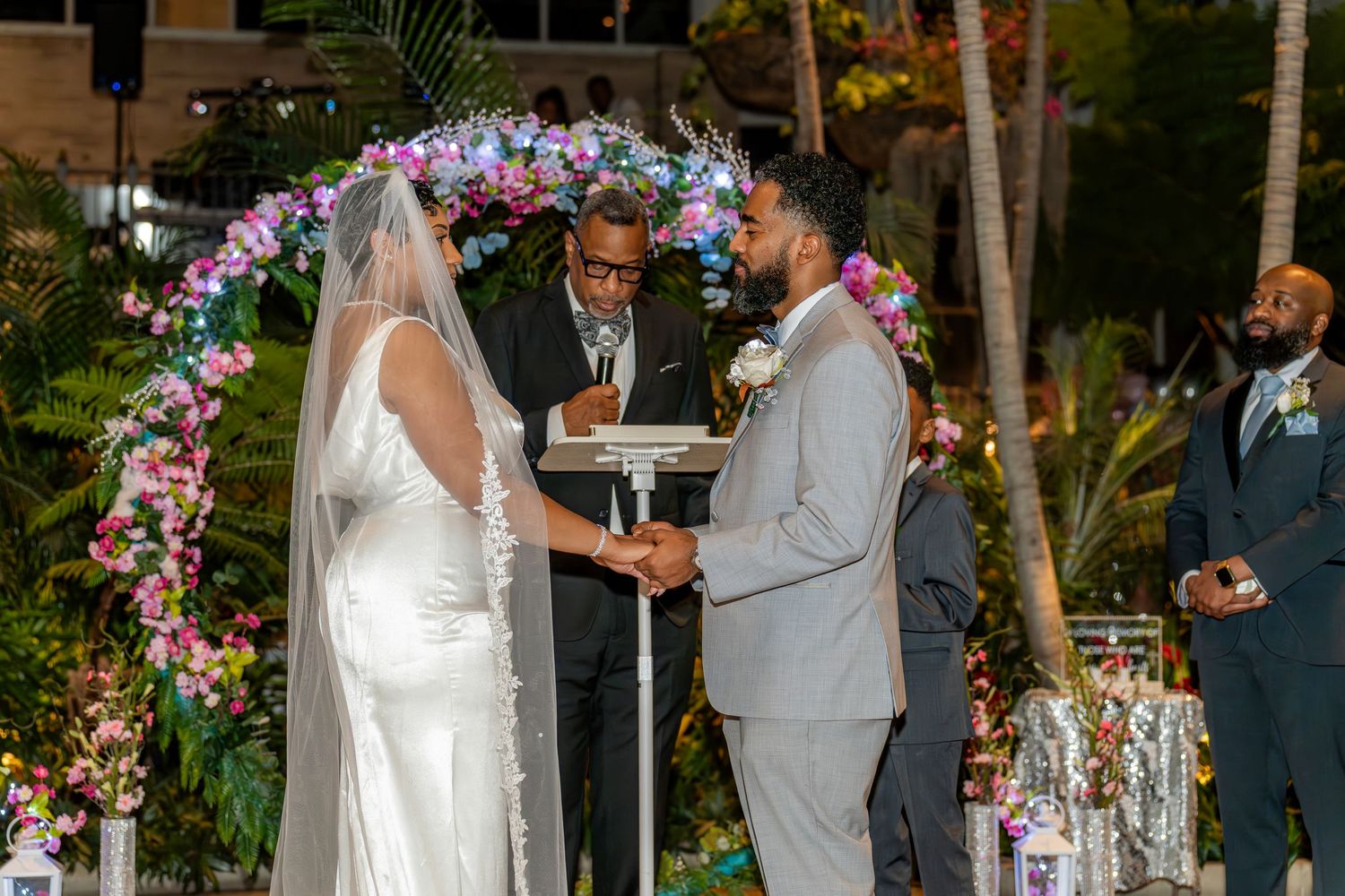 Couple holds hands before officiant under floral arch with purple flowers at outdoor night wedding.