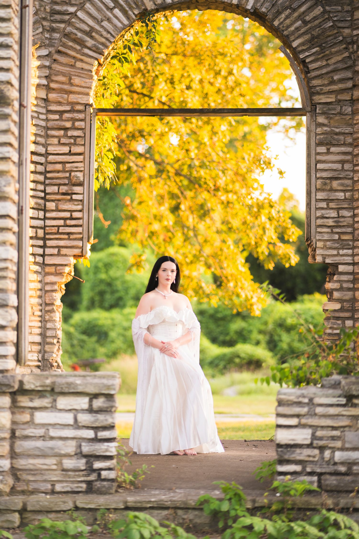 Pregnant woman in flowing white dress standing in stone archway surrounded by autumn foliage.