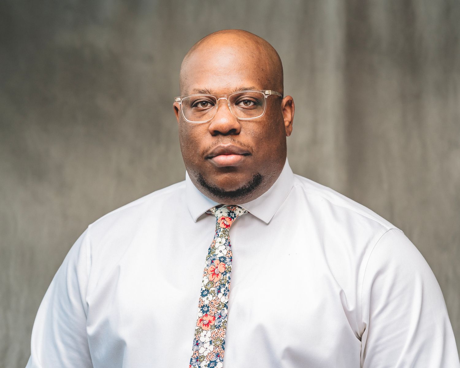 Professional headshot of a man in a white dress shirt and floral tie wearing clear-framed glasses.