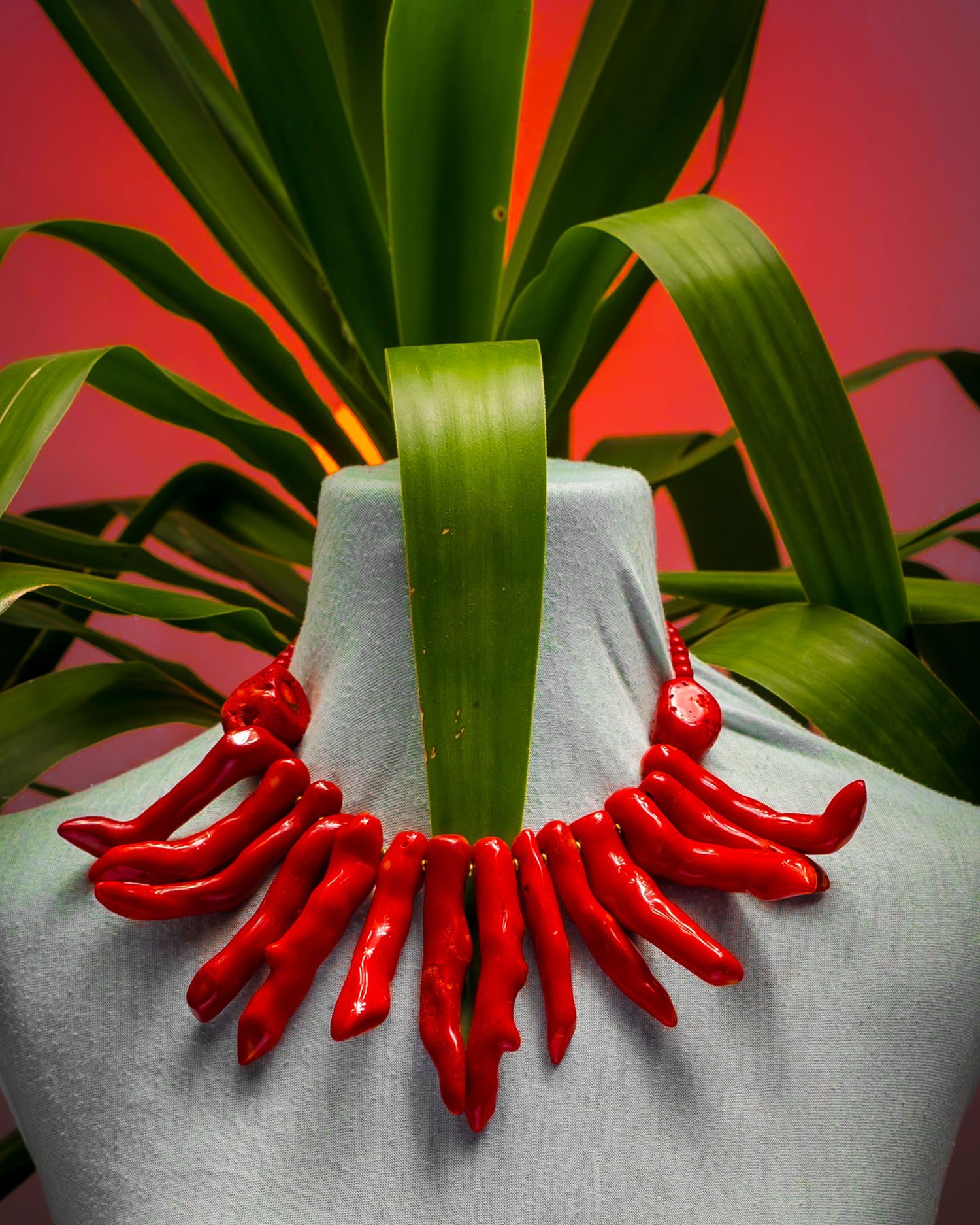 Bold red coral branch statement necklace displayed on gray mannequin with tropical plant backdrop.