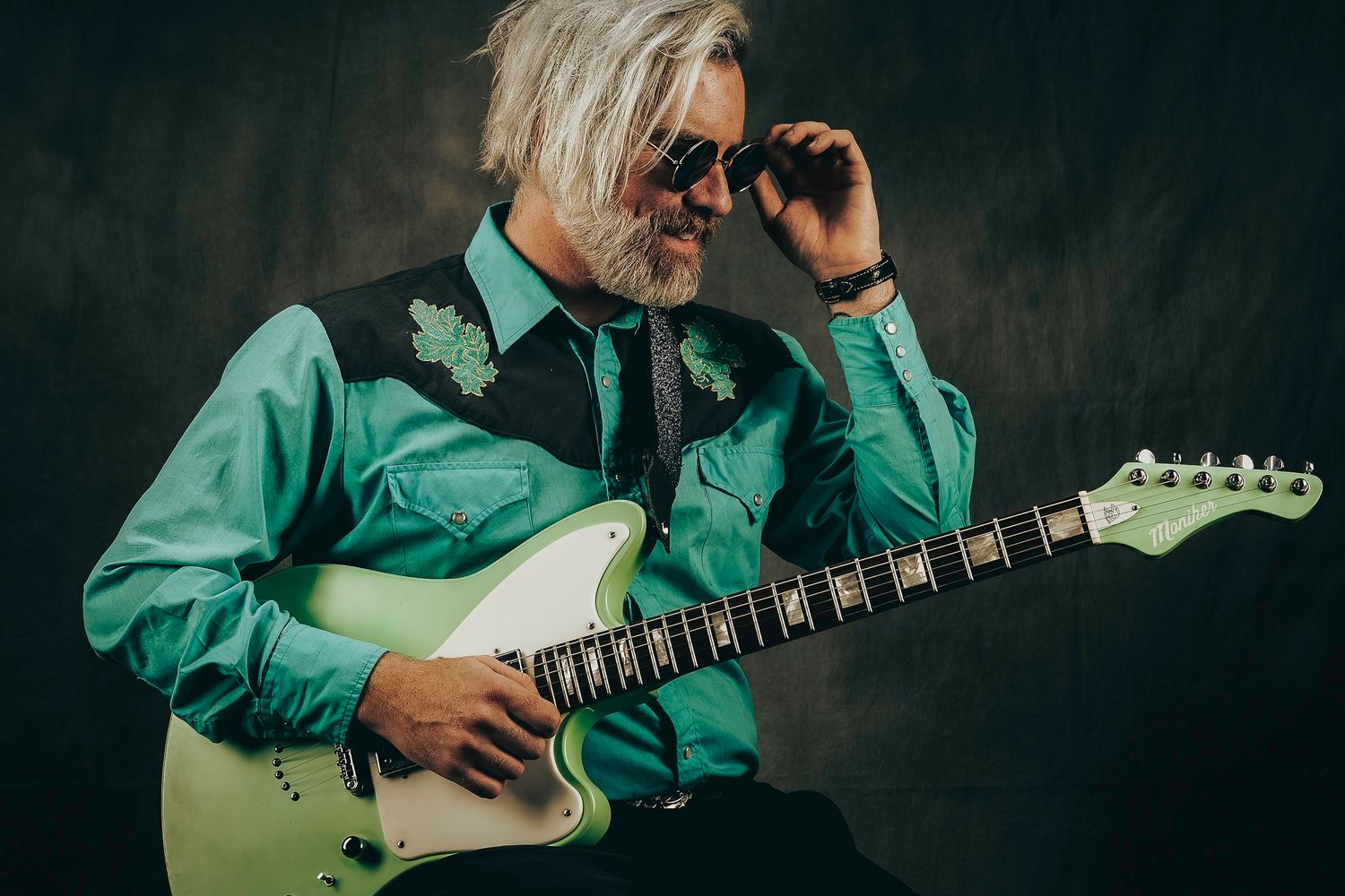 Older musician with white hair plays a mint green electric guitar wearing a teal western shirt.