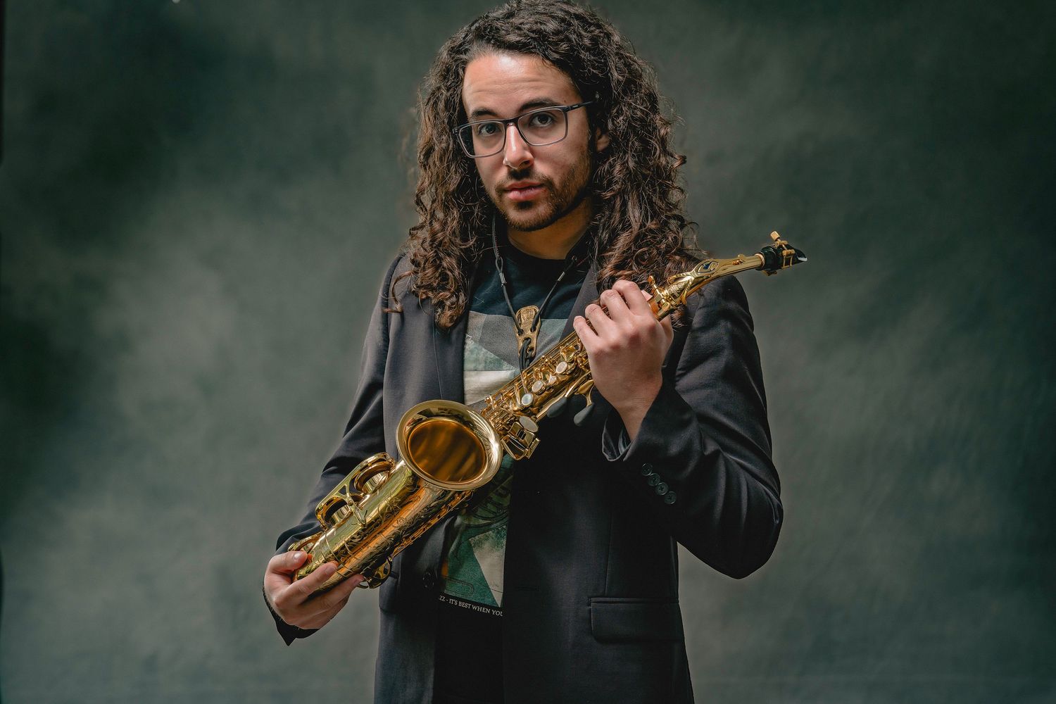 Curly-haired saxophonist in a black blazer holds a gold alto saxophone in a dramatic studio portrait.