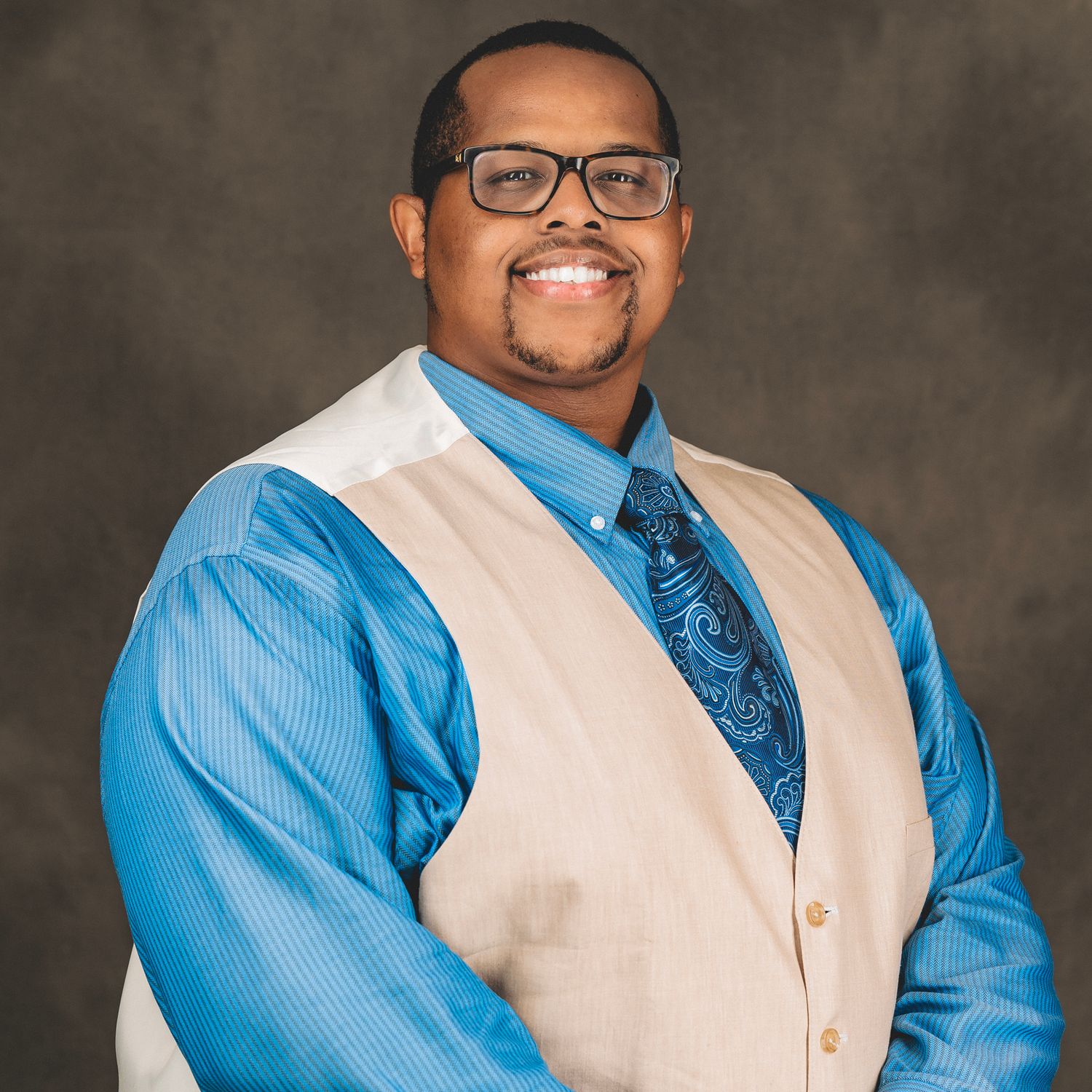 Smiling man wearing glasses, a blue shirt, and beige vest in a professional portrait.