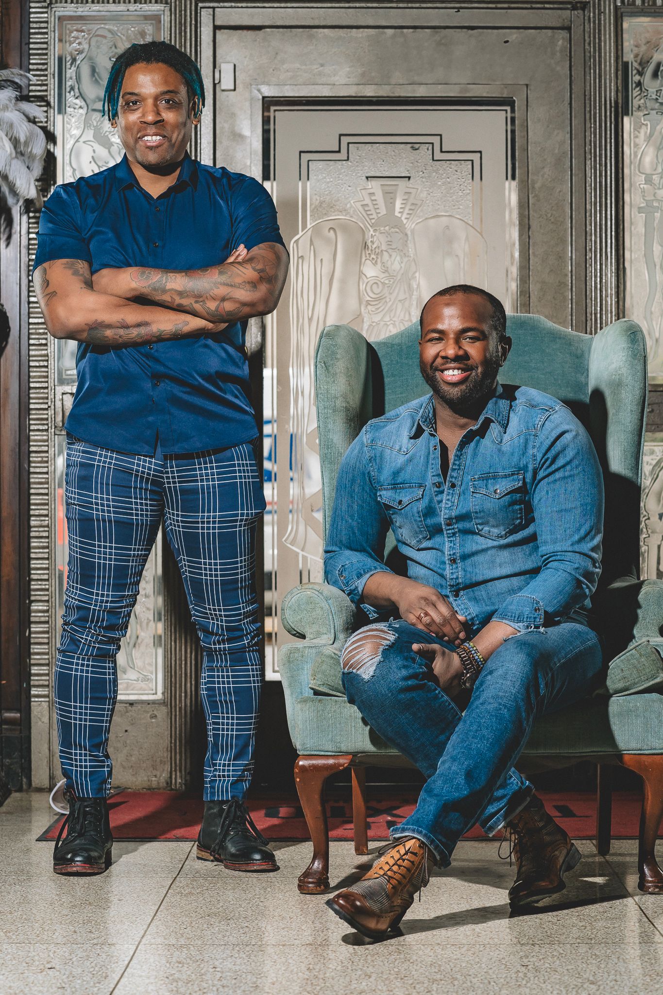 Two stylish men posing in elegant interior, one standing and one seated in teal armchair.