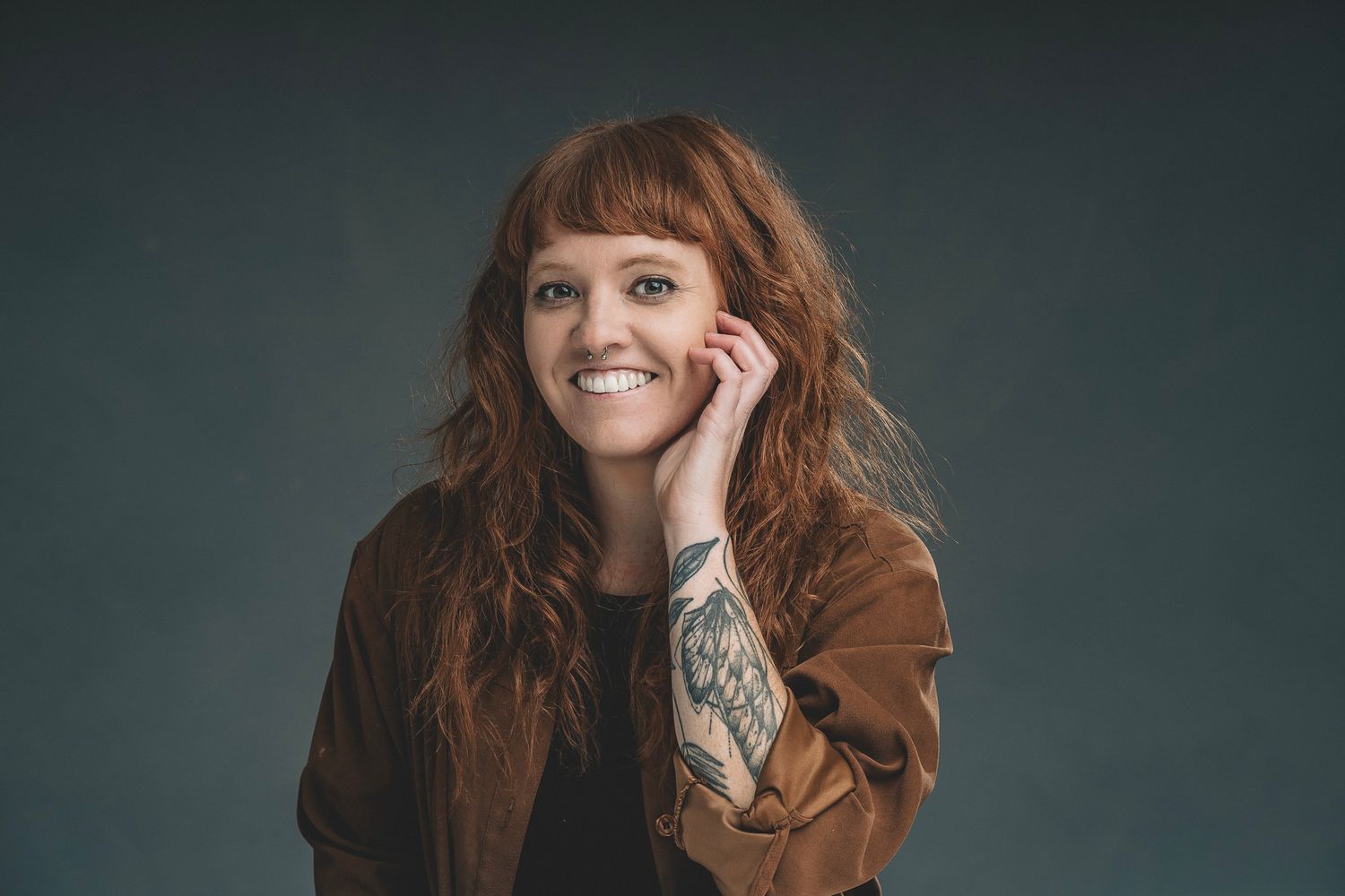 Smiling woman with long red hair and tattoo wearing a brown jacket in a studio portrait.