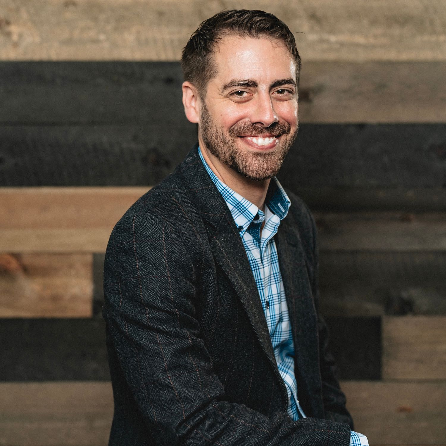 Smiling man with beard wearing a dark blazer and plaid shirt in a professional headshot.