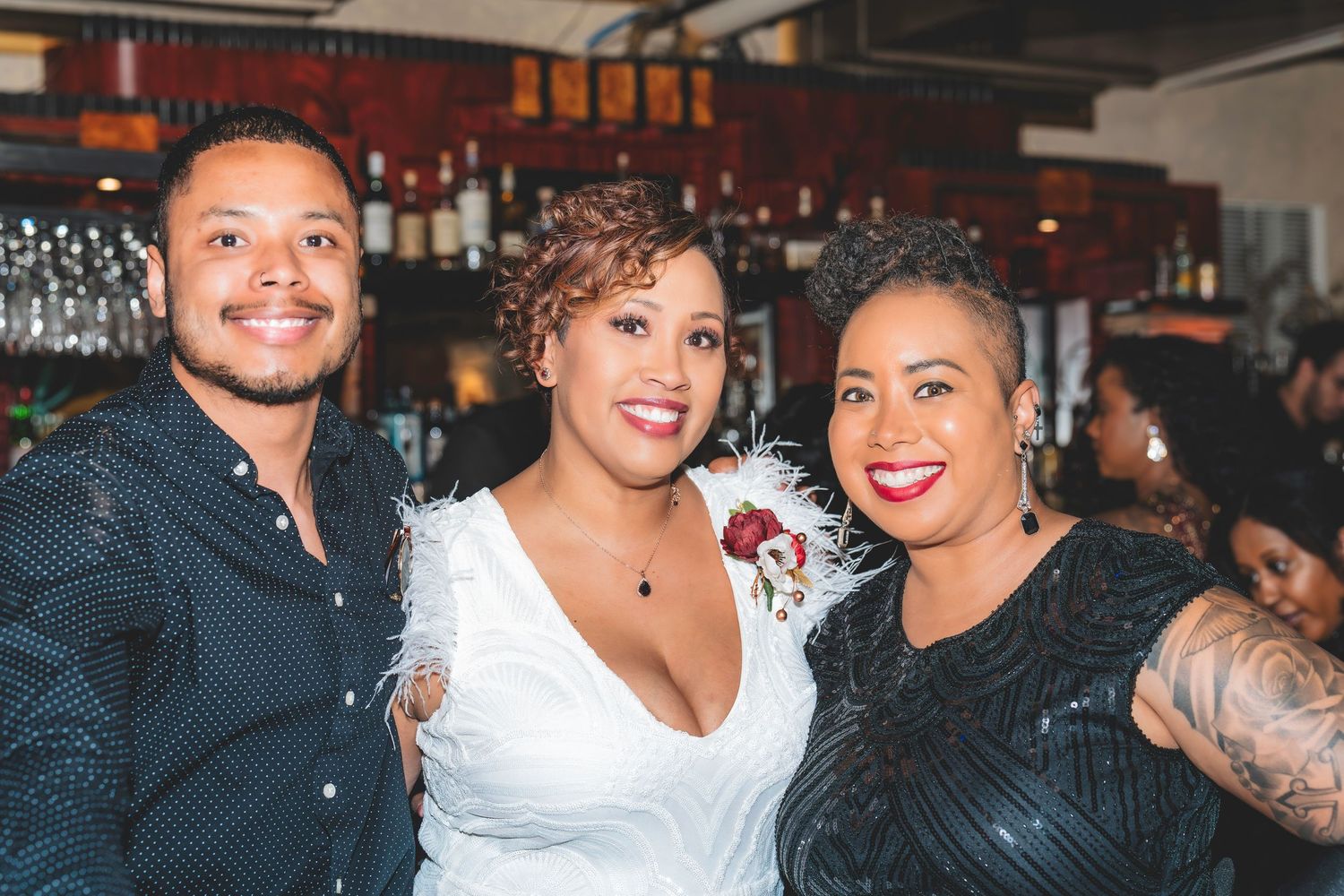 Bride in white feathered dress smiles with two guests at a lively wedding reception bar.