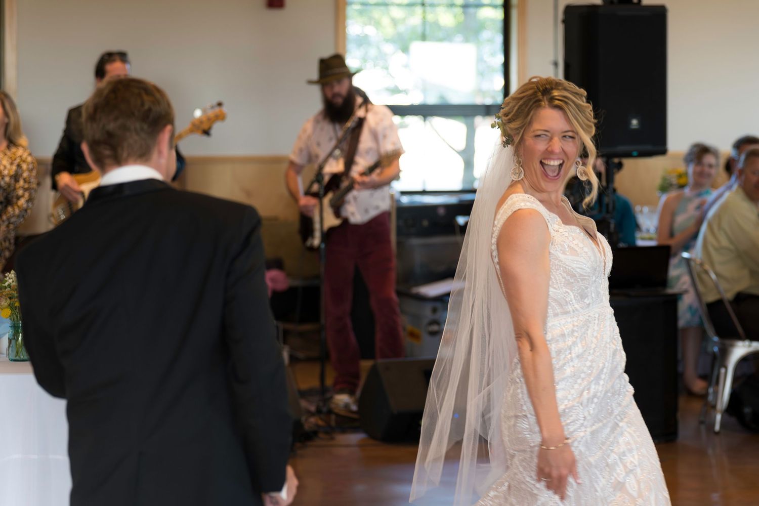 Bride laughing with veil flowing during first dance while live band plays at wedding reception.