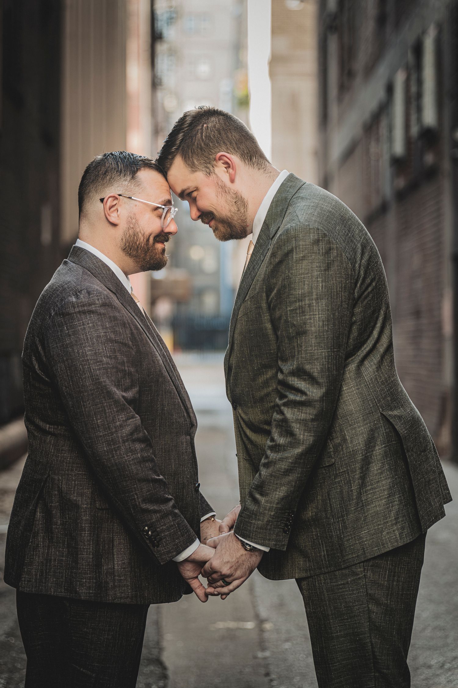 Two grooms in gray suits hold hands and touch foreheads in a romantic alley wedding portrait.