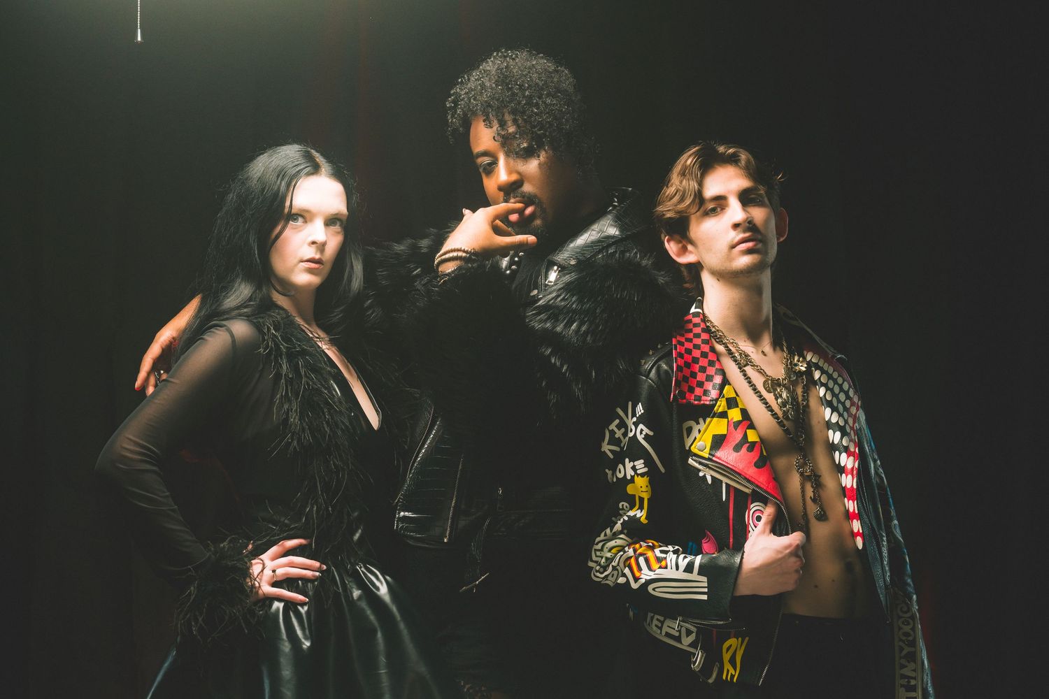 Three edgy fashion models pose dramatically in dark clothing against a black background.